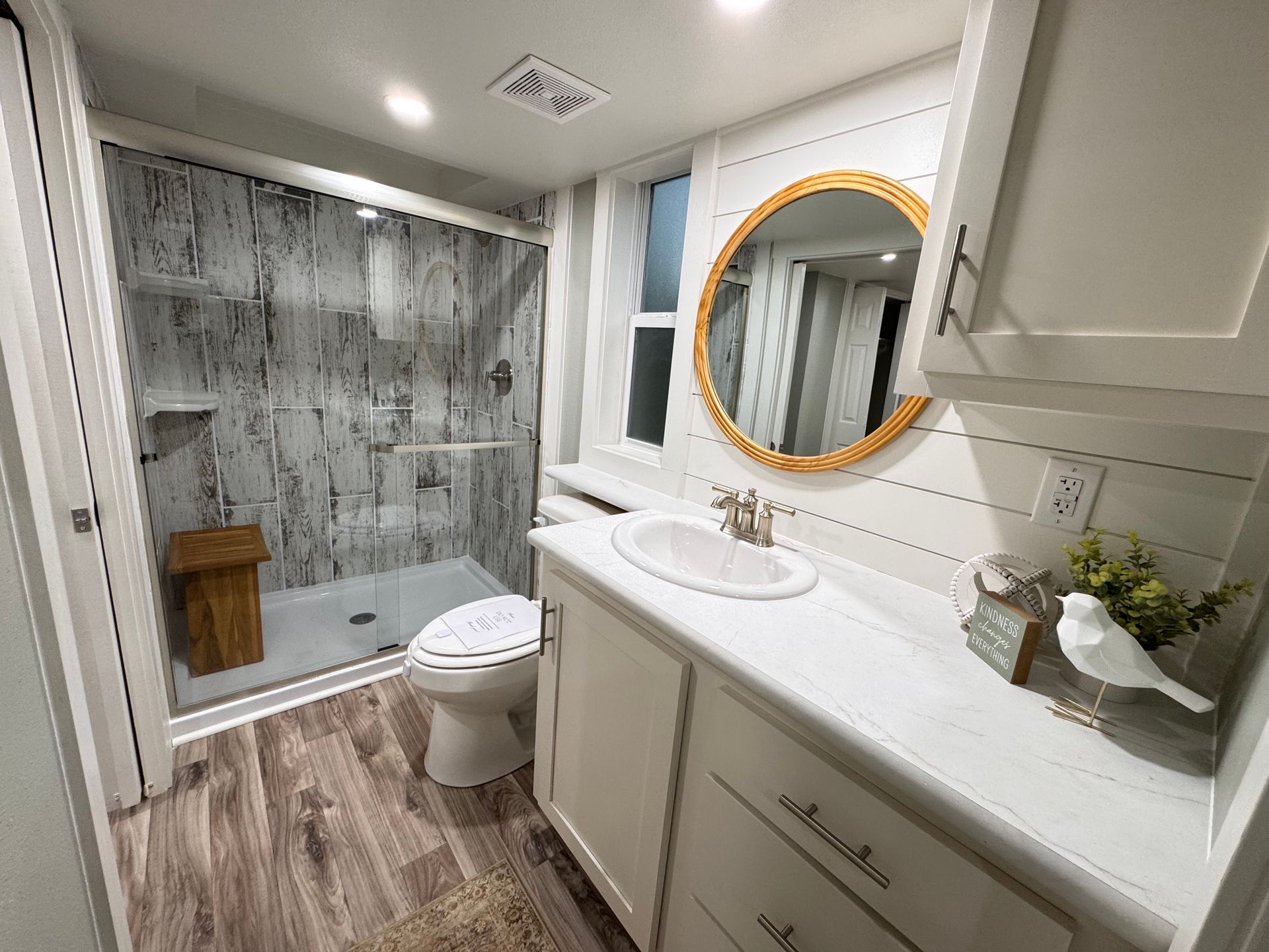 A bathroom with white shiplap walls, a wooden-framed mirror, a white vanity, a walk-in shower, and wood-look flooring.