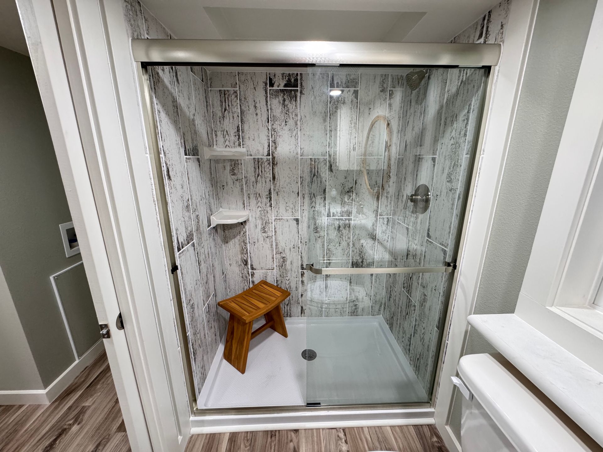 A walk-in shower with white and grey marble-patterned walls, a glass sliding door, and a wooden bench inside.
