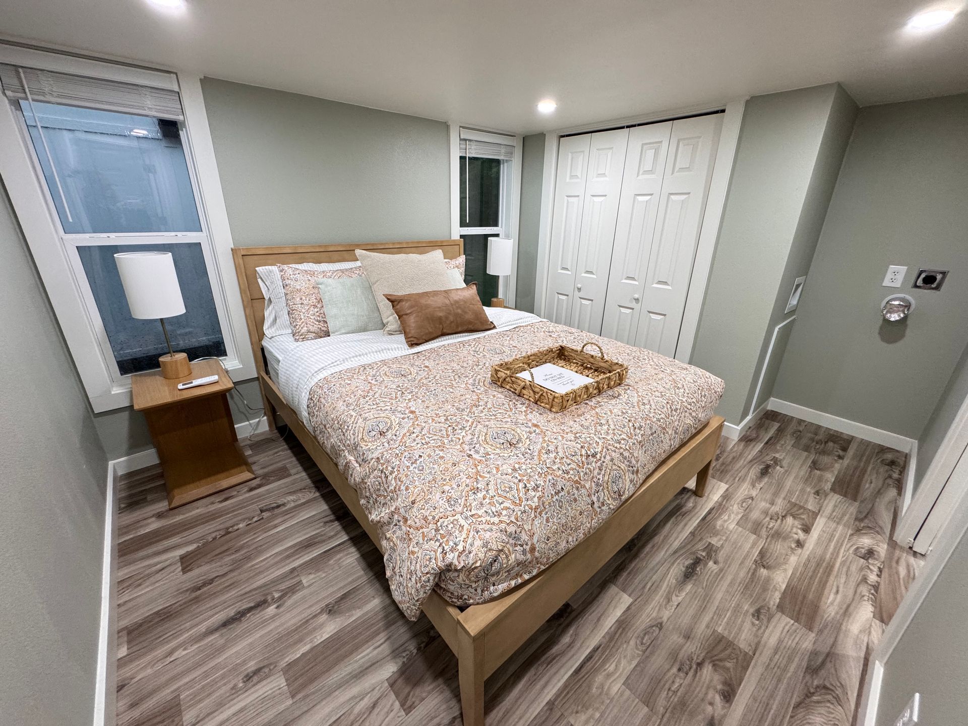 A cozy bedroom featuring a wooden bed with floral bedding, a side table, lamp, and grey walls with light wood flooring.
