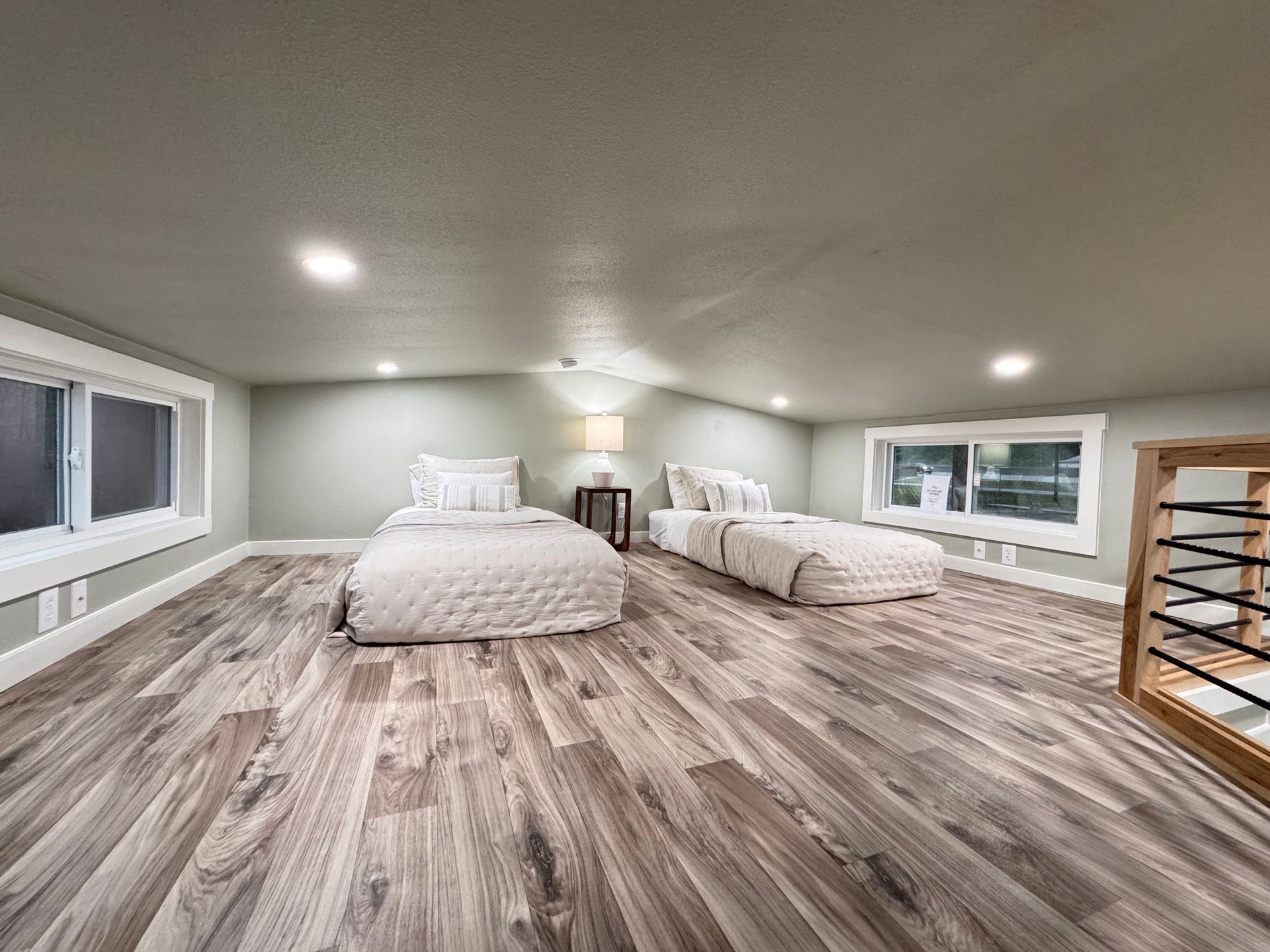 An attic bedroom with a sloped ceiling, two single beds with white bedding, wood-look flooring, and two windows.