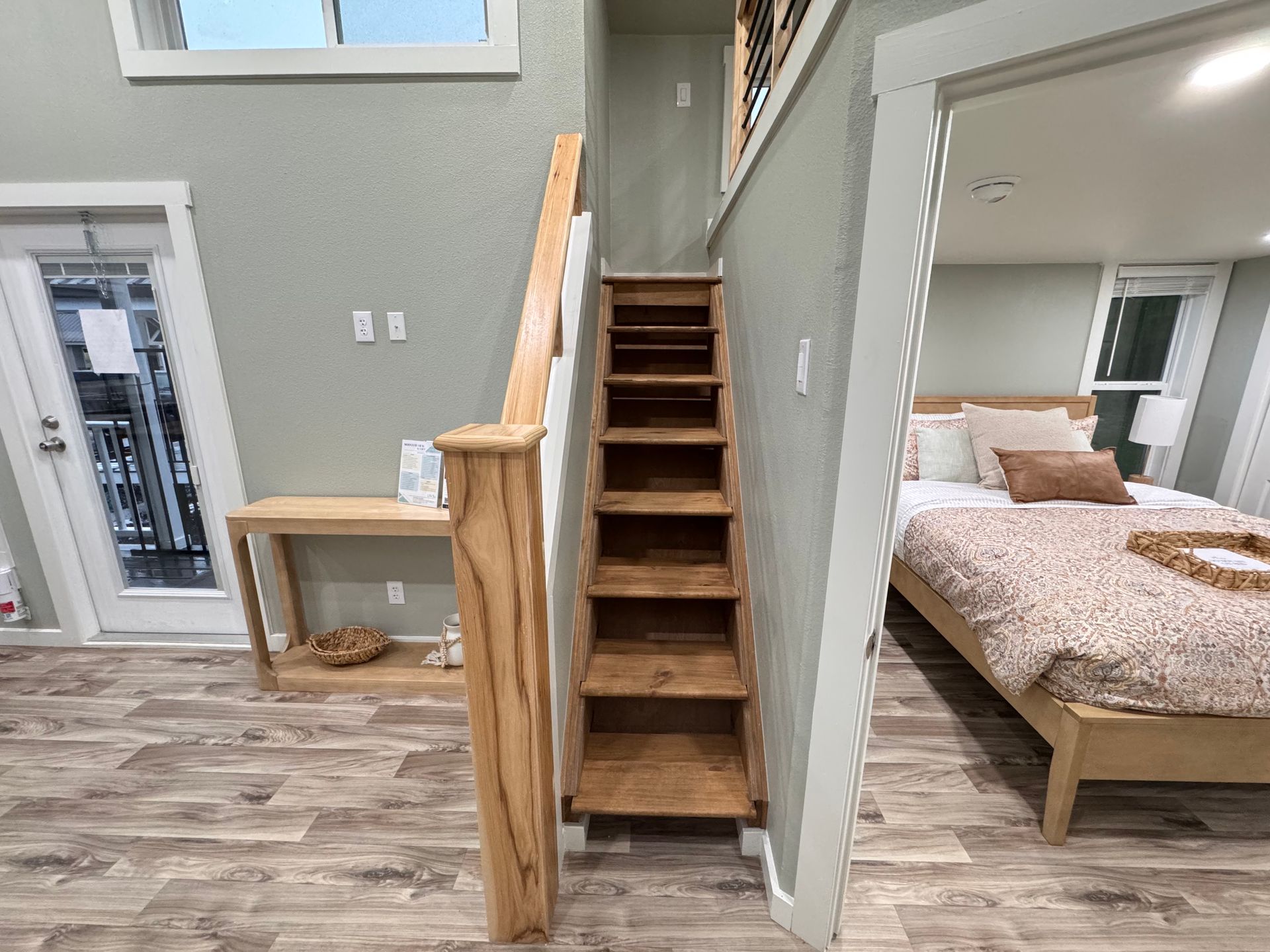 A set of wooden stairs leading to a loft, located between a hallway console table and a bedroom with a made bed.
