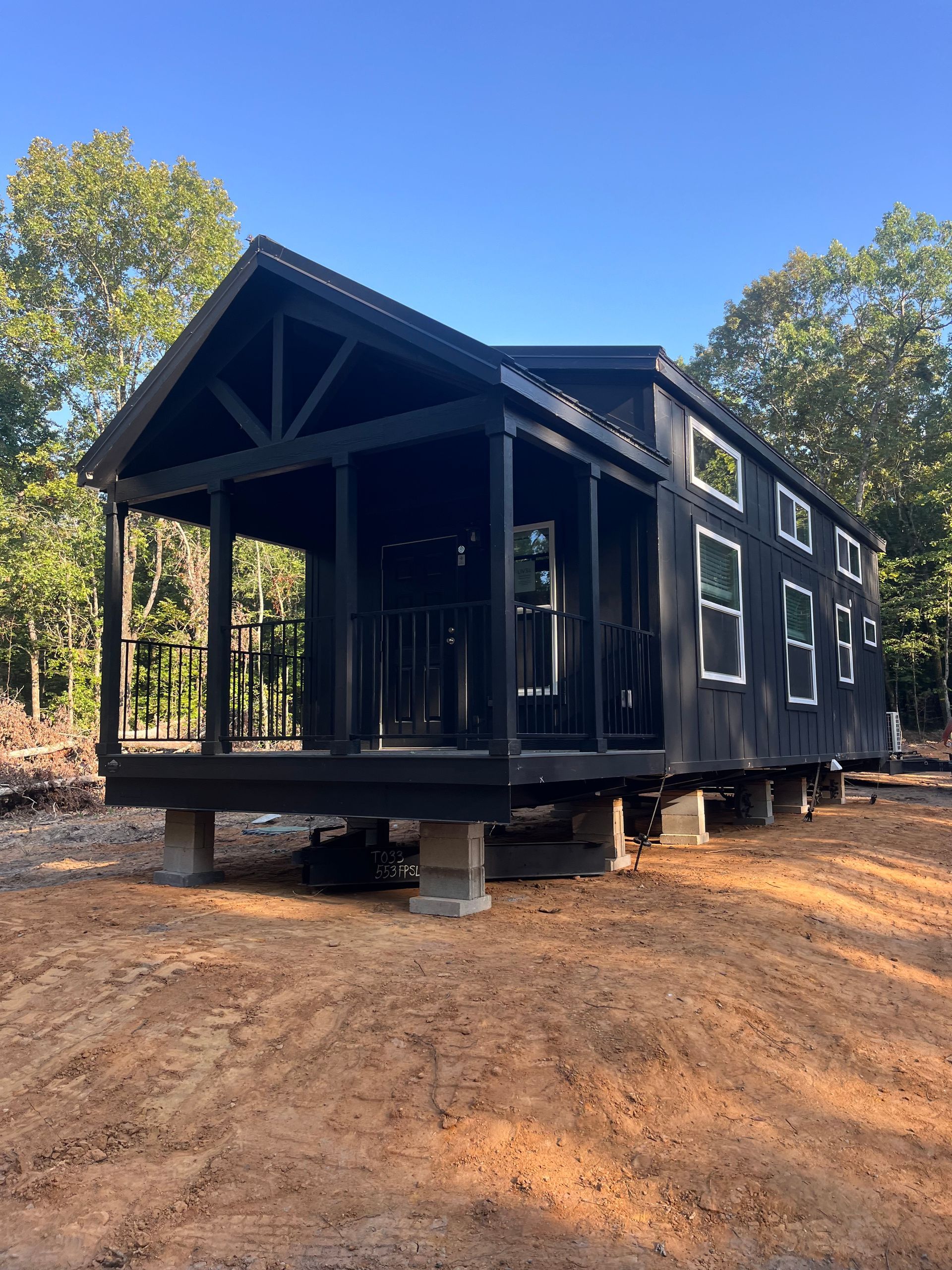 Black tiny house with a porch, supported by concrete blocks, in a wooded area.