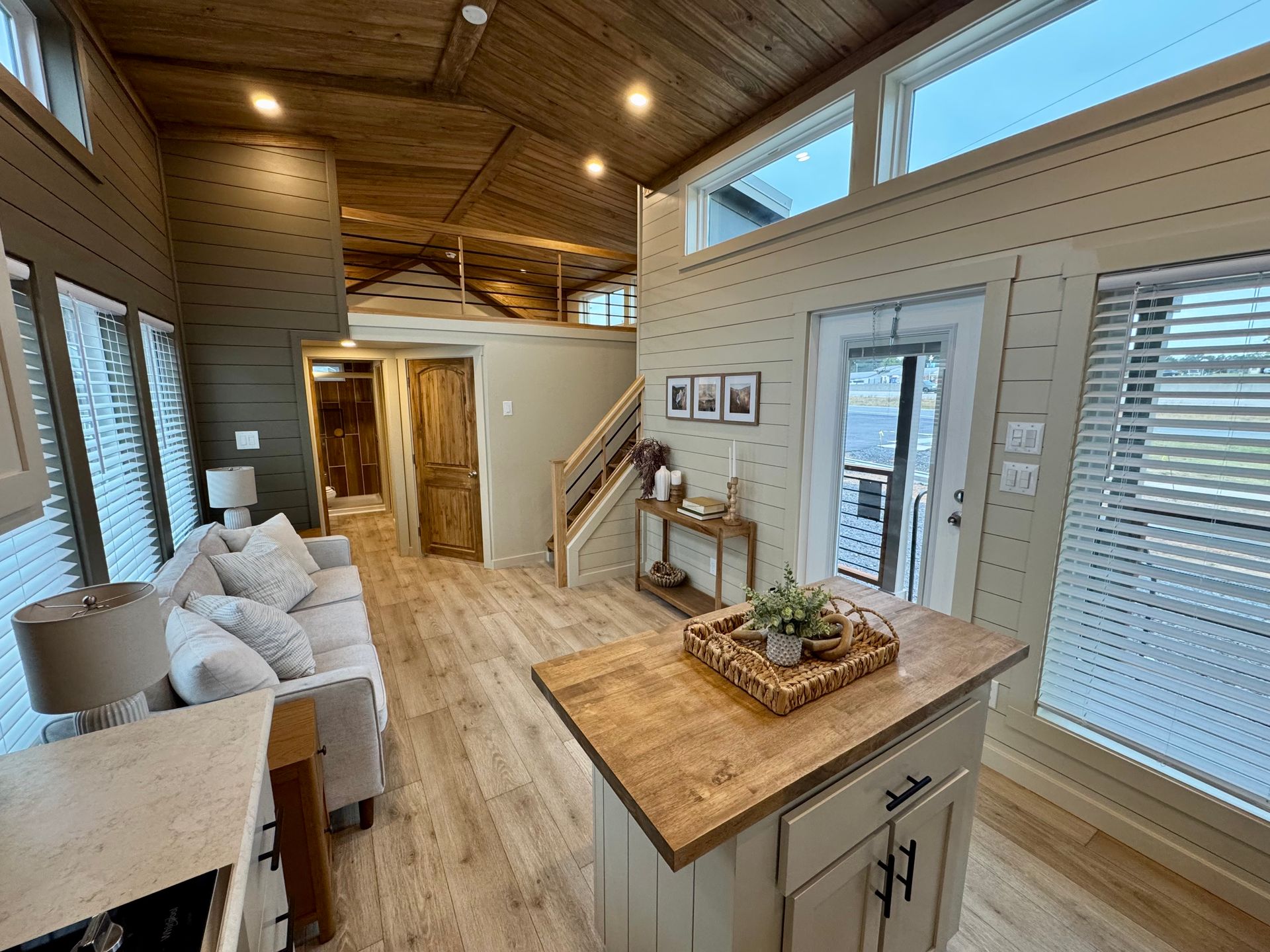 A light-filled tiny house interior with a neutral sofa, wooden kitchen island, and stairs leading to a sleeping loft.