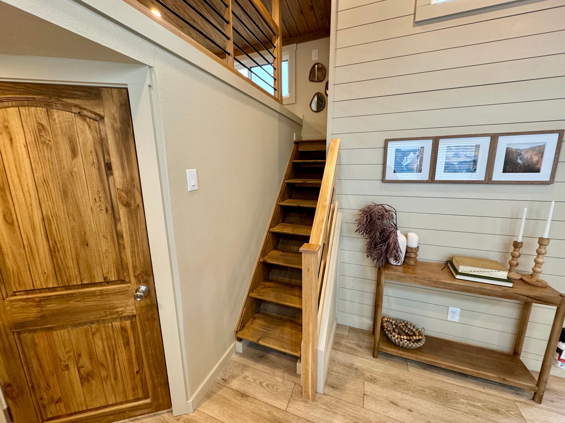 A wooden staircase leads to a loft beside a wooden door and a console table with three framed pictures on a shiplap wall.