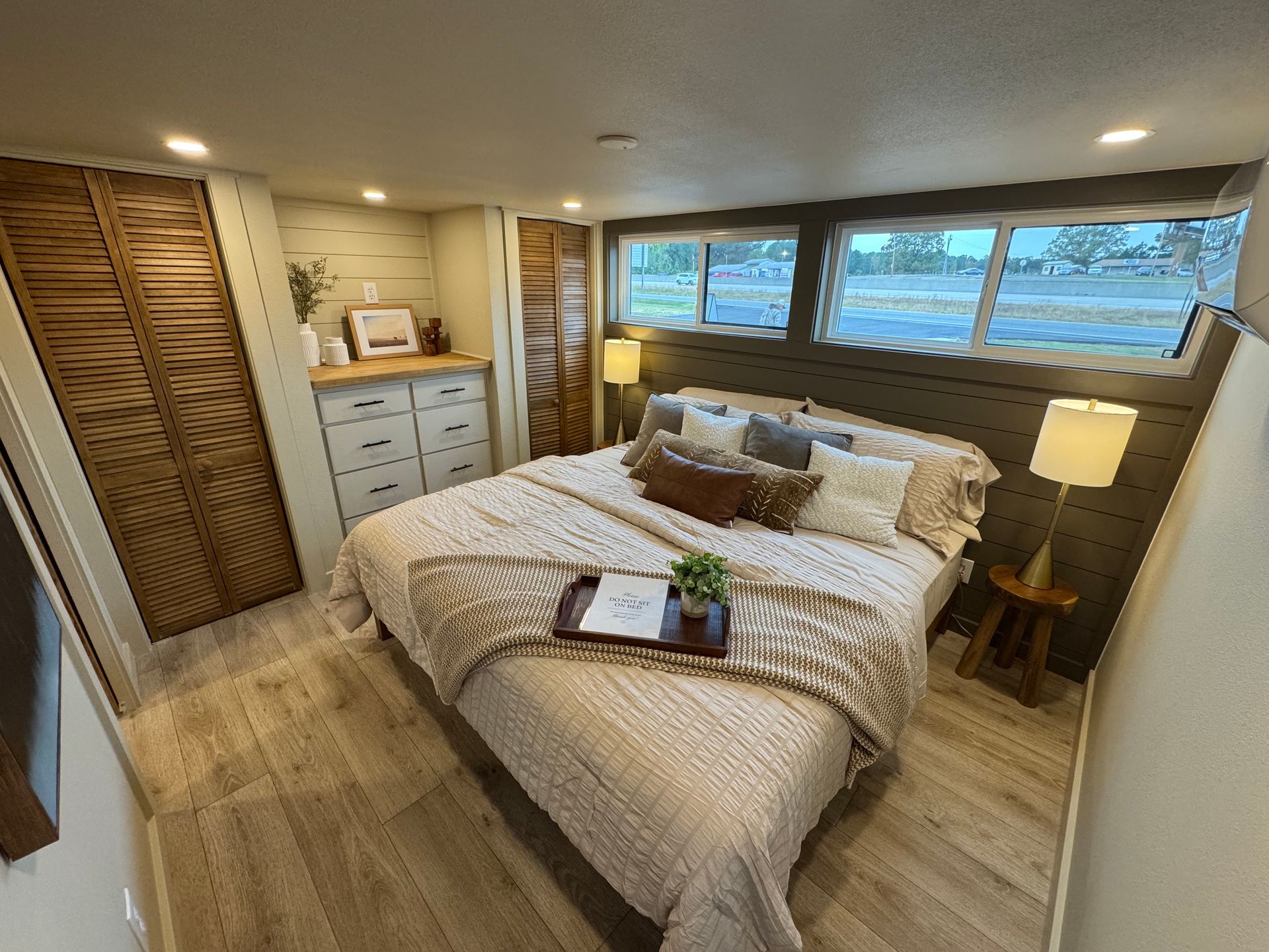 A cozy bedroom with a queen bed, wooden flooring, louvered closet doors, and windows looking out to a field.