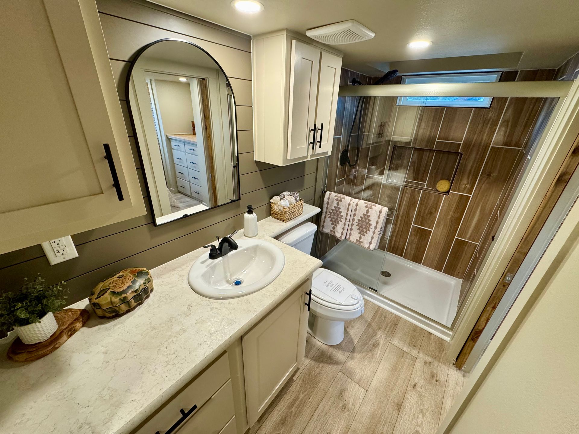 A bright bathroom featuring a white vanity with a rounded sink, an arched mirror, a toilet, and a glass-enclosed shower.