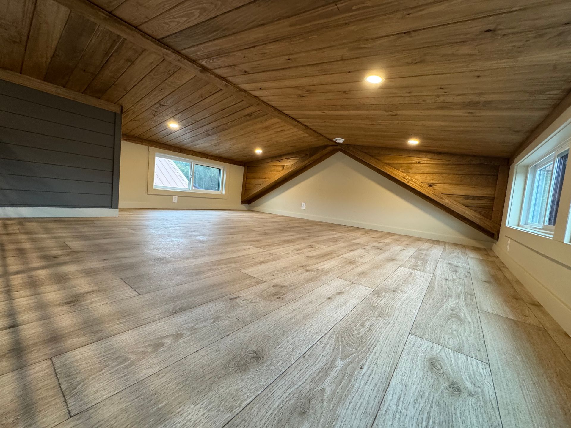 An interior loft with wood-paneled sloped ceilings, light wood-look flooring, and windows on both ends.