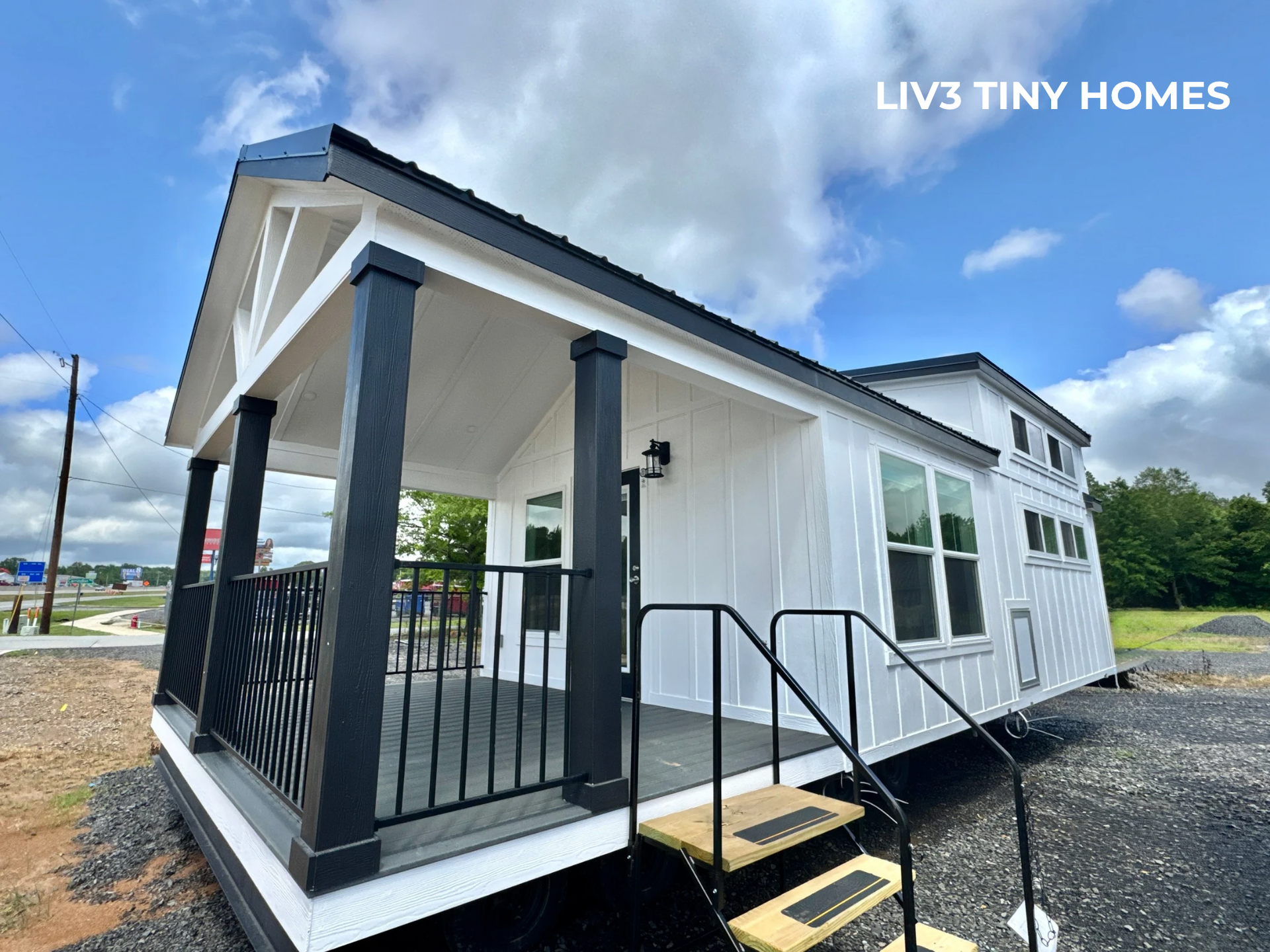 A white tiny home with a black-railed porch and staircase stands on a gravel lot under a cloudy blue sky.
