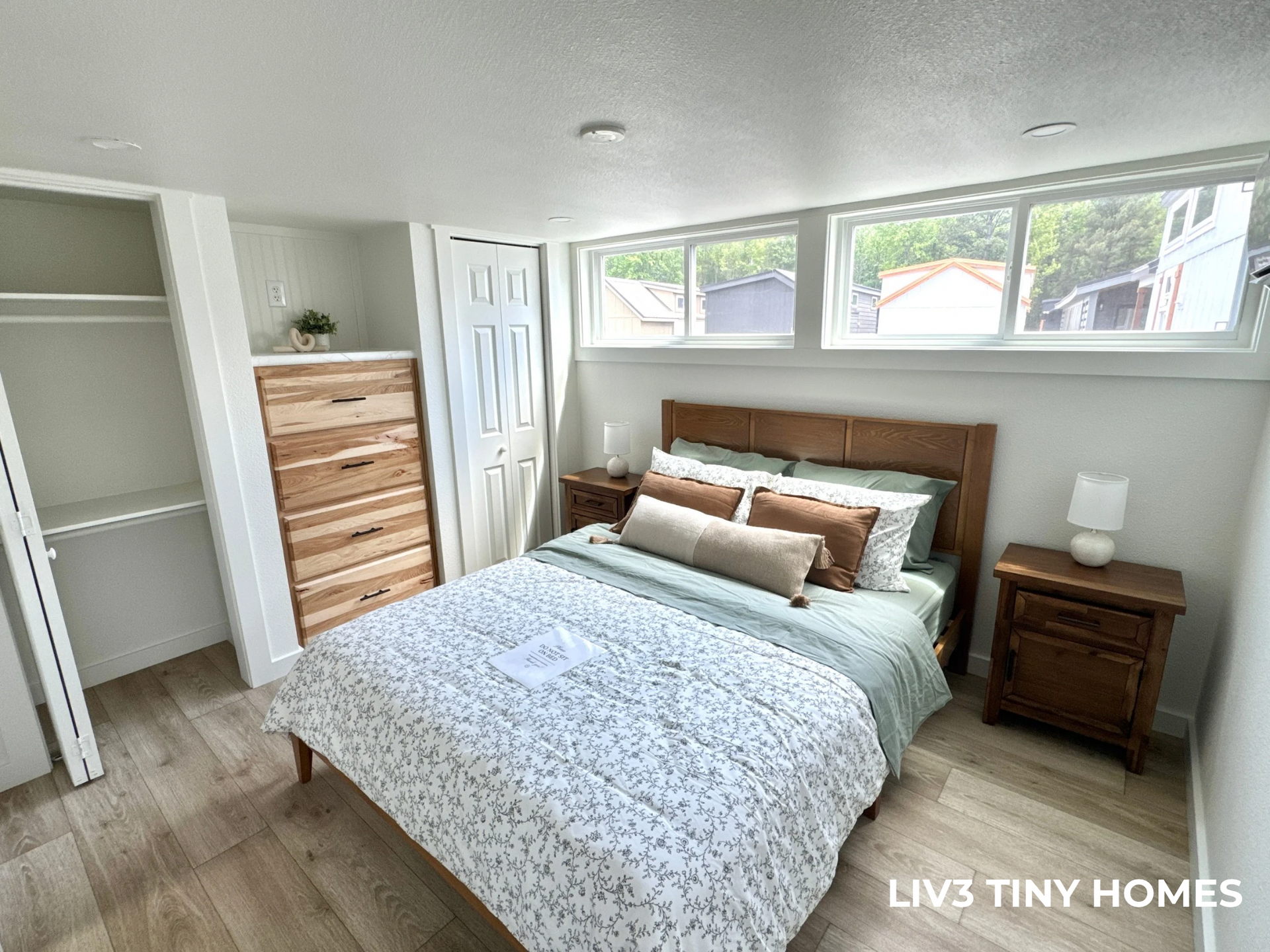 A bedroom in a tiny home with a bed, nightstand, wooden dresser, closet, and horizontal windows along the wall.