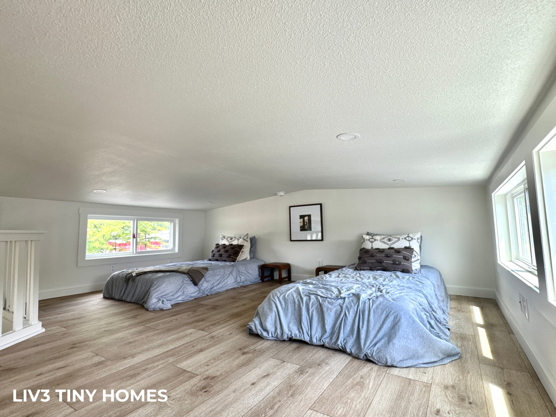 A bedroom loft with two twin beds featuring grey bedding, light wood floors, white walls, and windows on both sides.