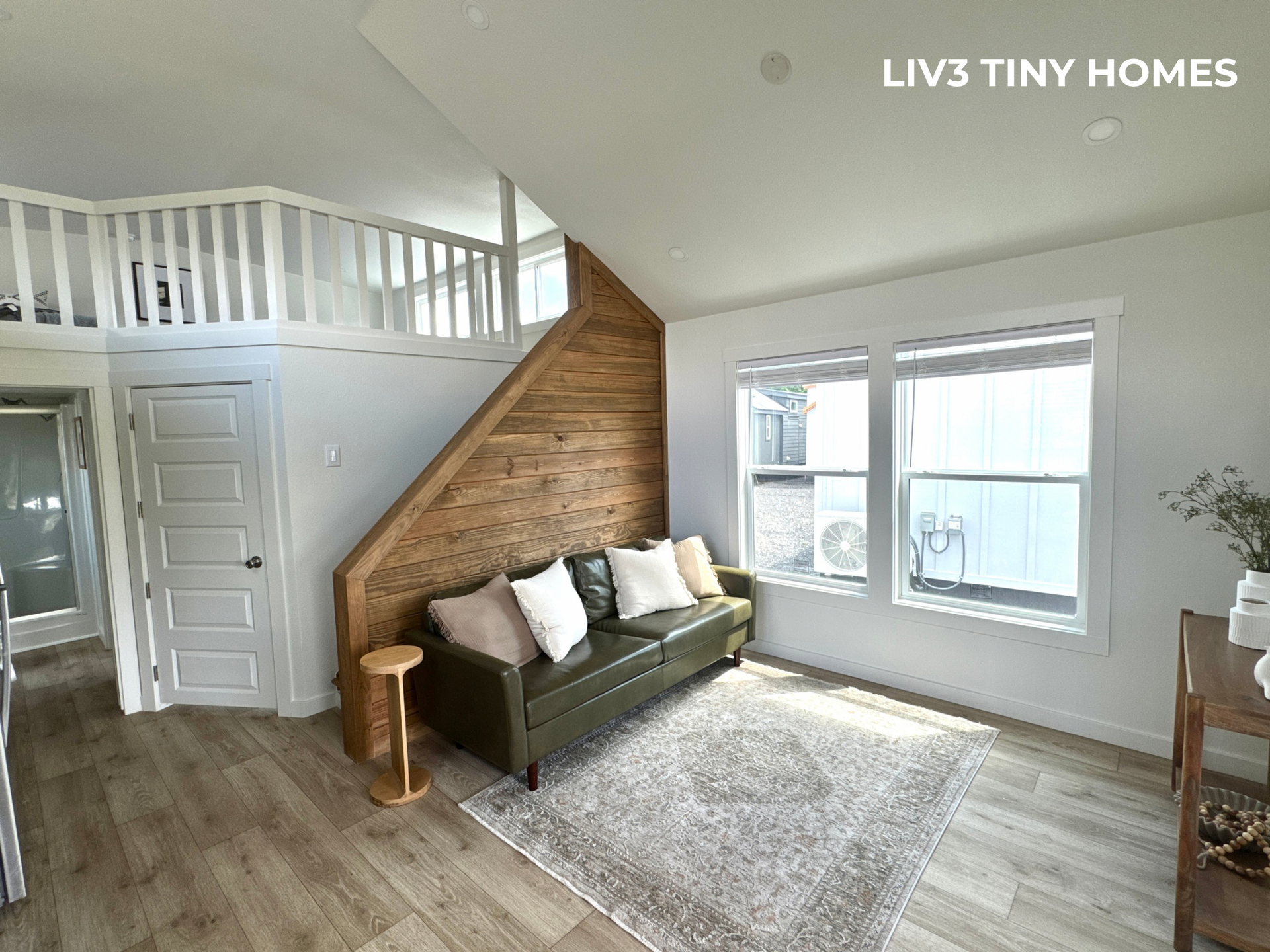 A cozy living area in a tiny home with a green leather sofa, wood-paneled wall, loft area, and light wood flooring.