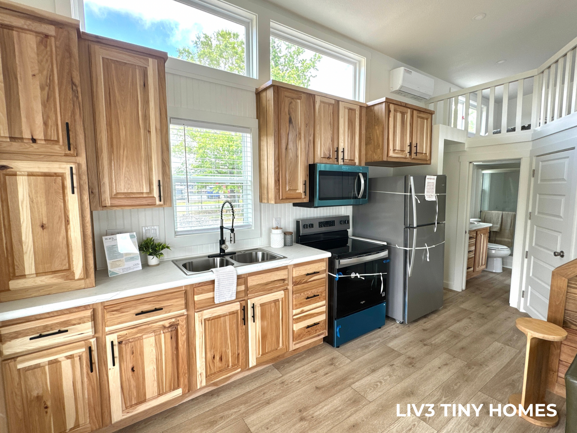 Modern tiny home kitchen with light wood cabinets, stainless steel appliances, and wood-look flooring.