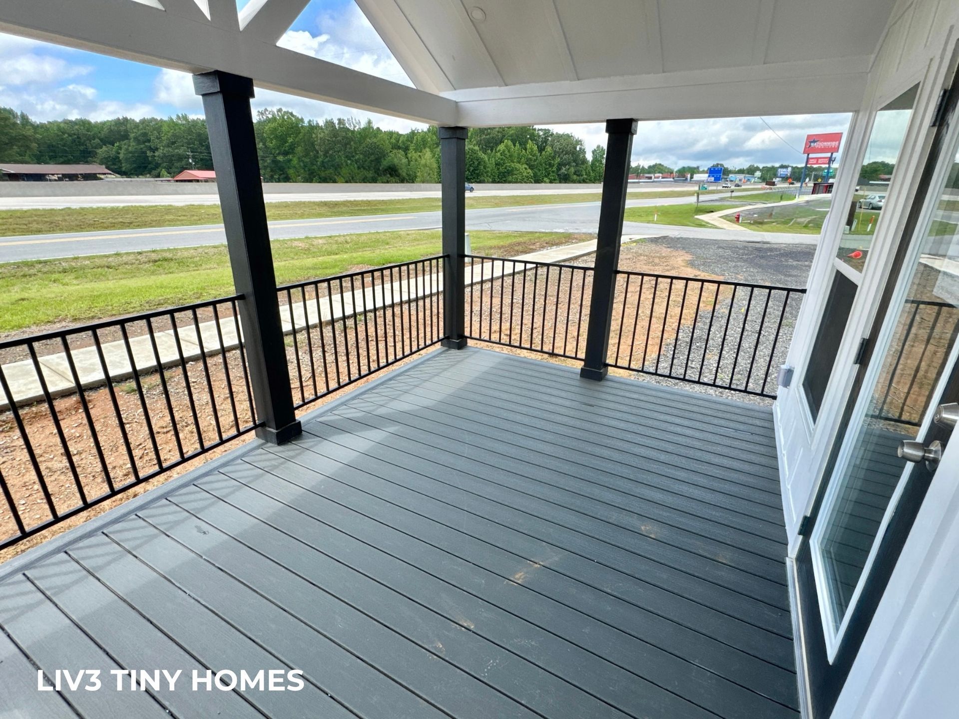 A covered porch with dark grey composite decking, black railings, and black support pillars, facing a grassy roadside.