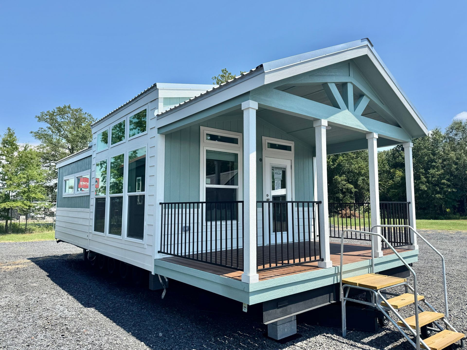 Tiny light blue house with porch, black railing, and steps on a gravel lot.