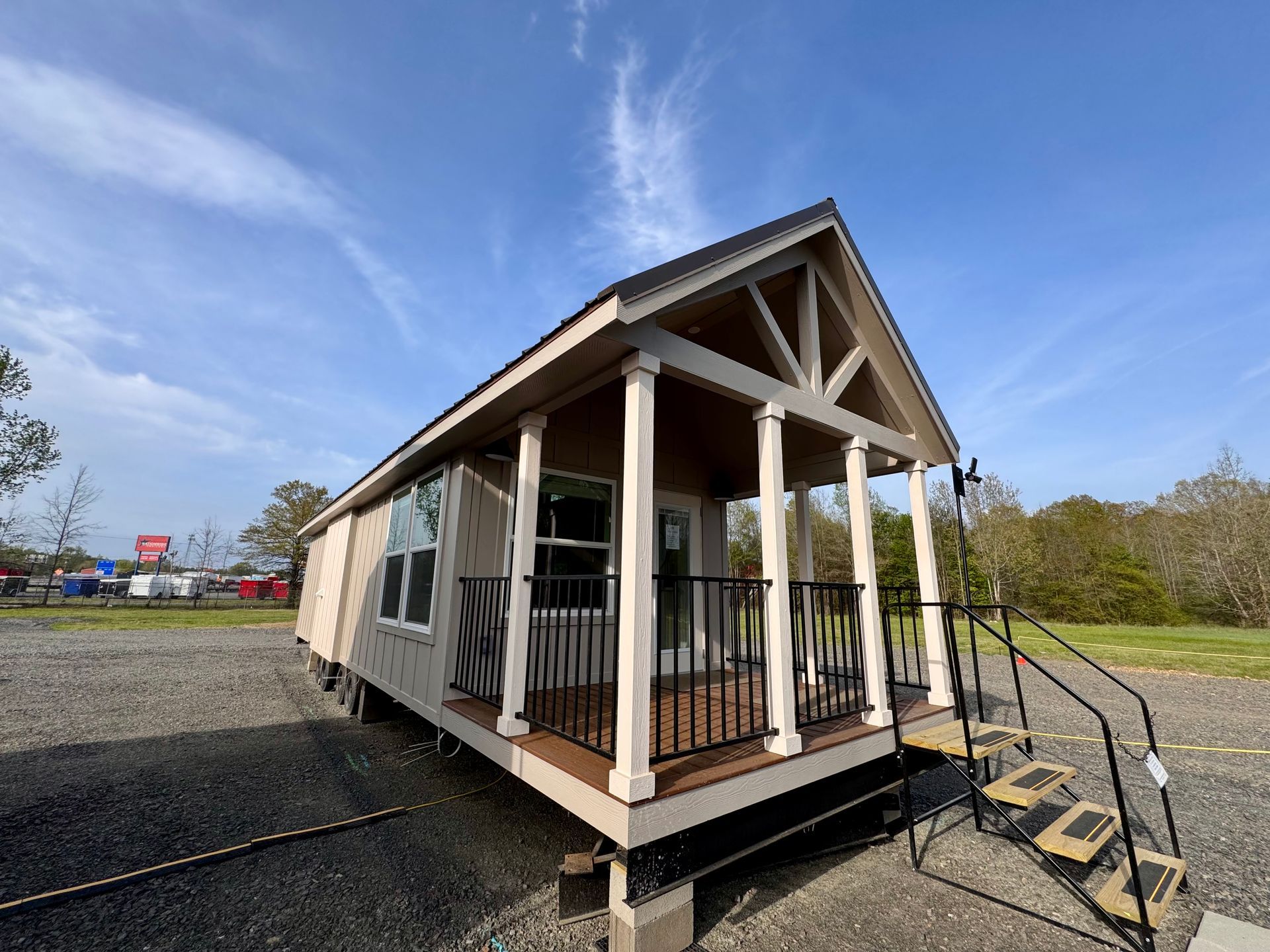 Small cabin on wheels with porch and stairs, tan and white exterior, blue sky background.