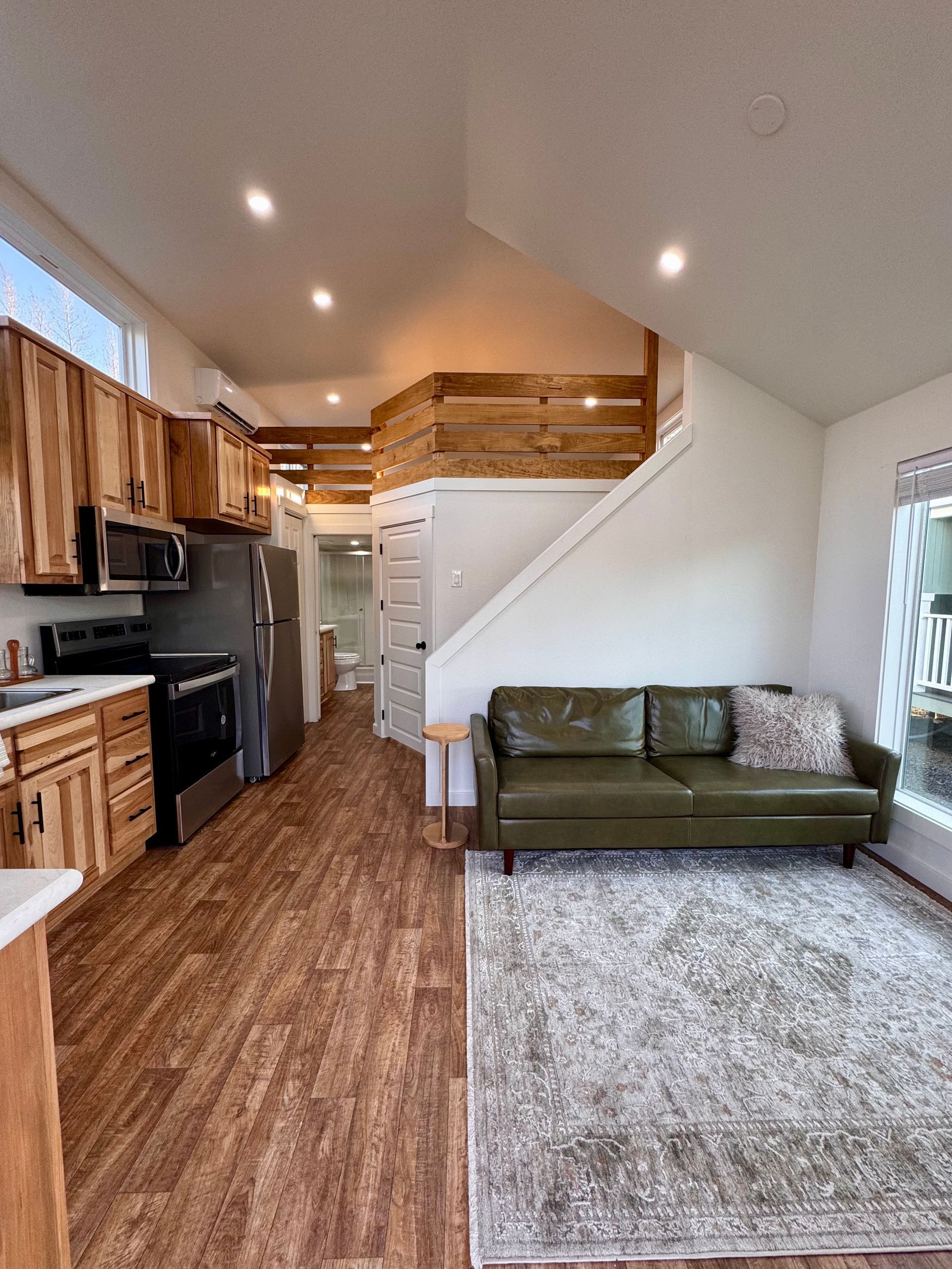 A small, open-concept living space with a kitchen, staircase to a loft, and a green leather sofa on a patterned rug.