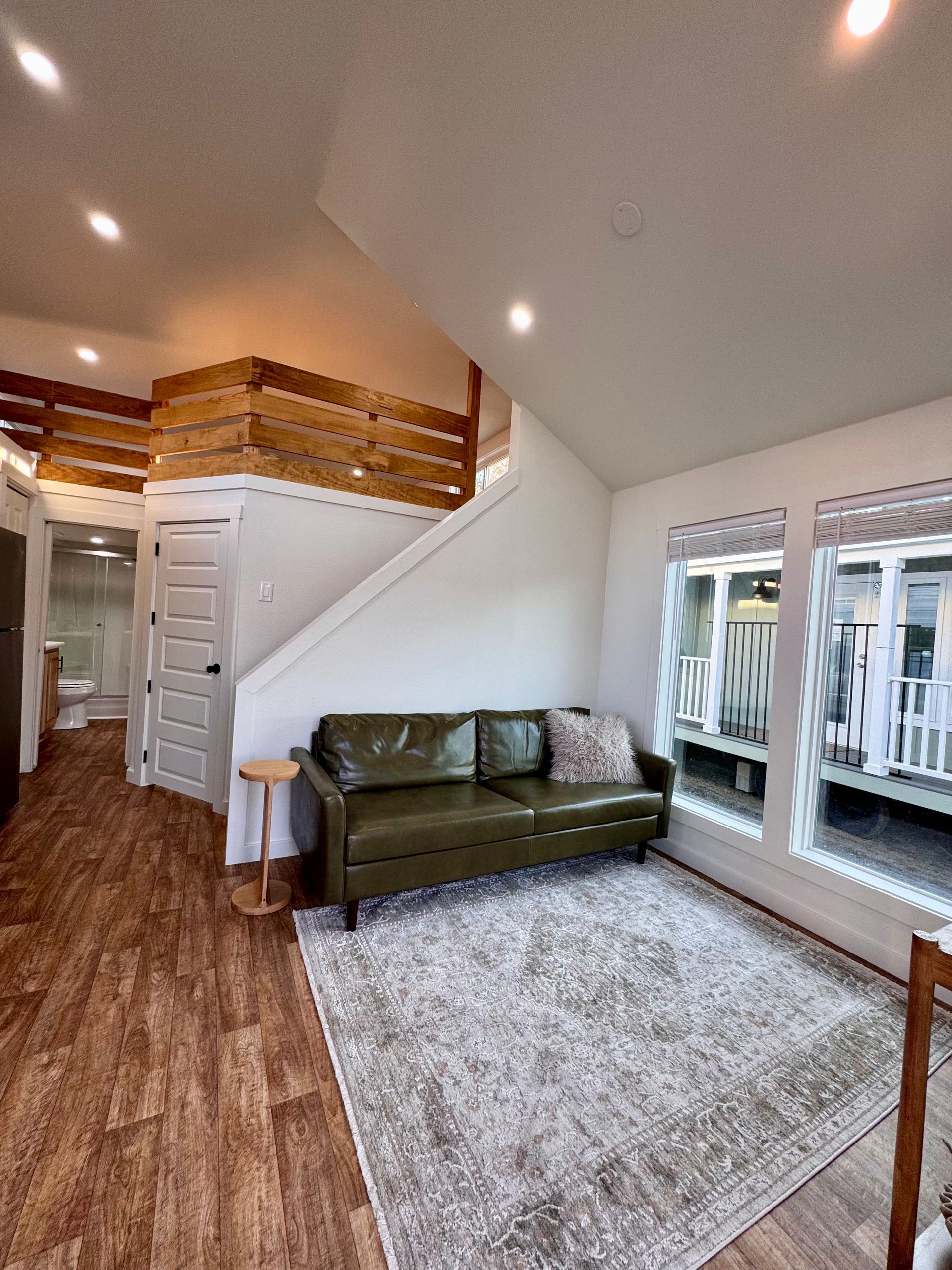 A modern living room with a green leather sofa on a large rug, wooden floors, a white staircase, and a wooden loft rail.