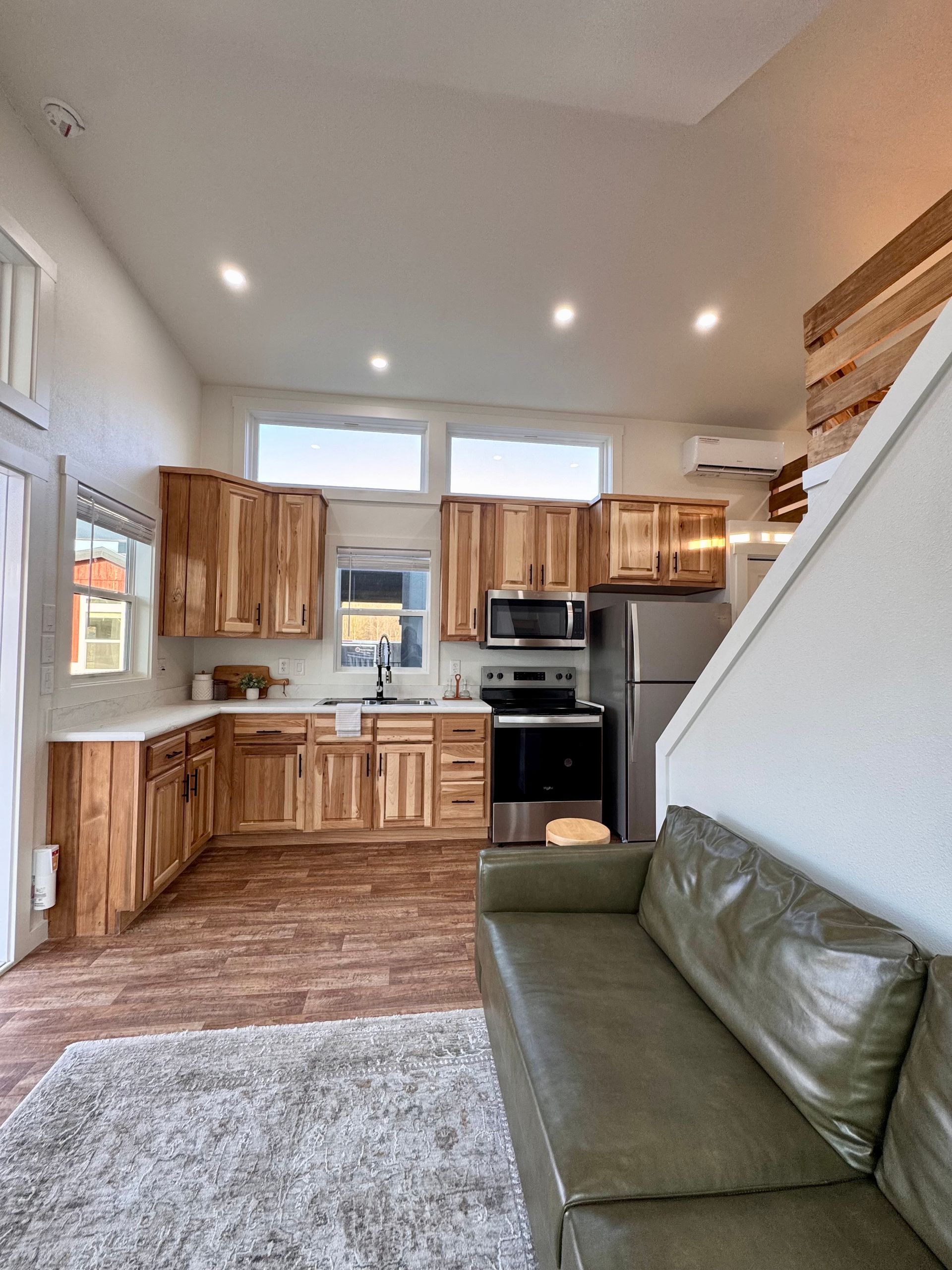 A modern kitchen with light wood cabinets and stainless steel appliances next to a green sofa on a textured area rug.