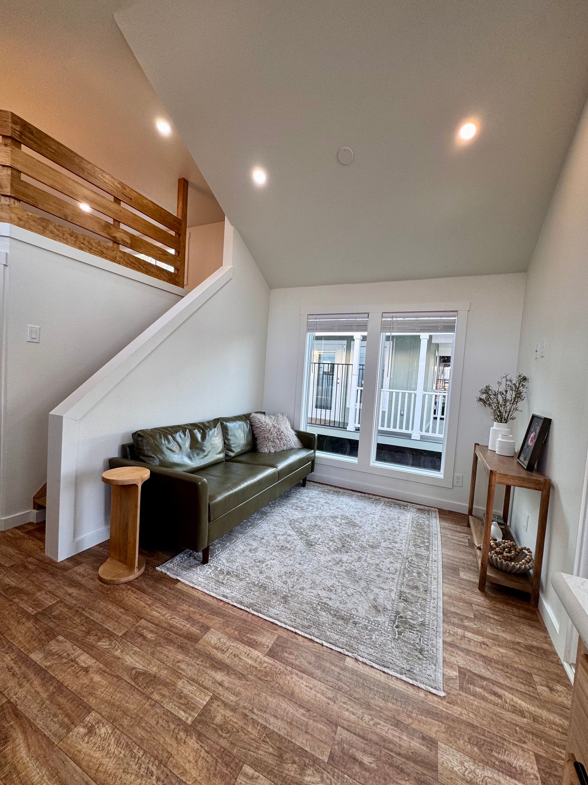 An olive green leather sofa sits on a grey area rug in a room with wood floors, a wooden staircase, and large windows.