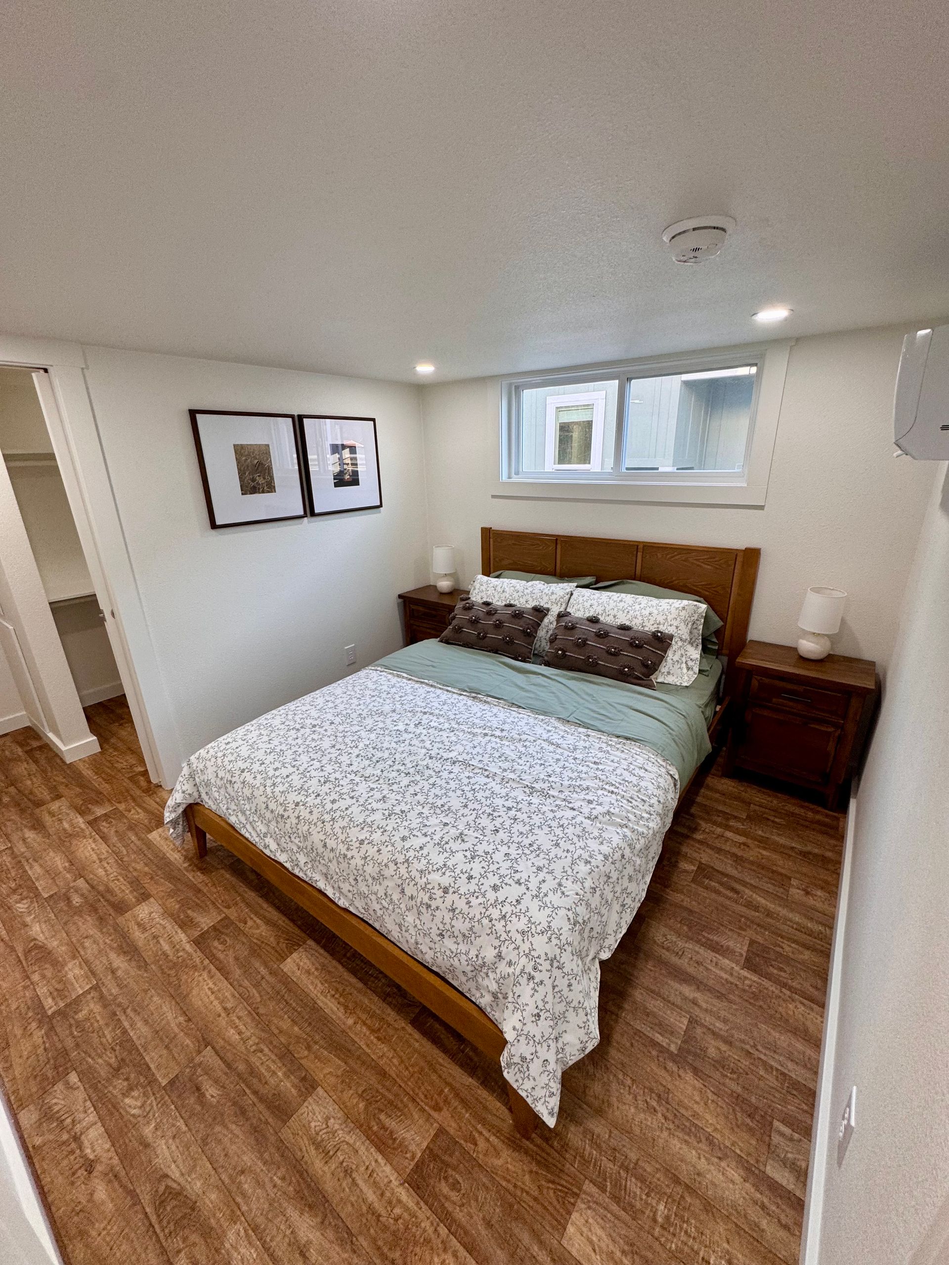 A bright bedroom with a queen bed, wooden headboard and nightstands, patterned bedding, two framed prints, and a window.