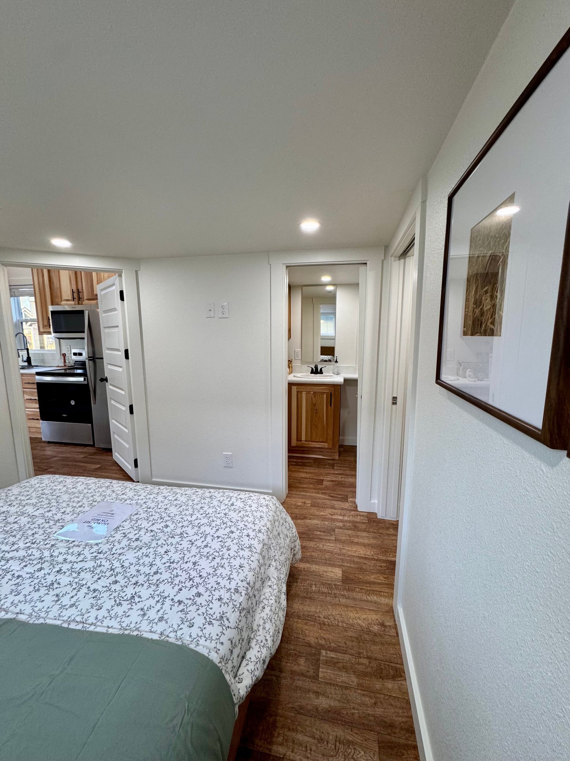 A bedroom view showing a bed with patterned bedding, a doorway to a kitchen, and another leading to a bathroom.