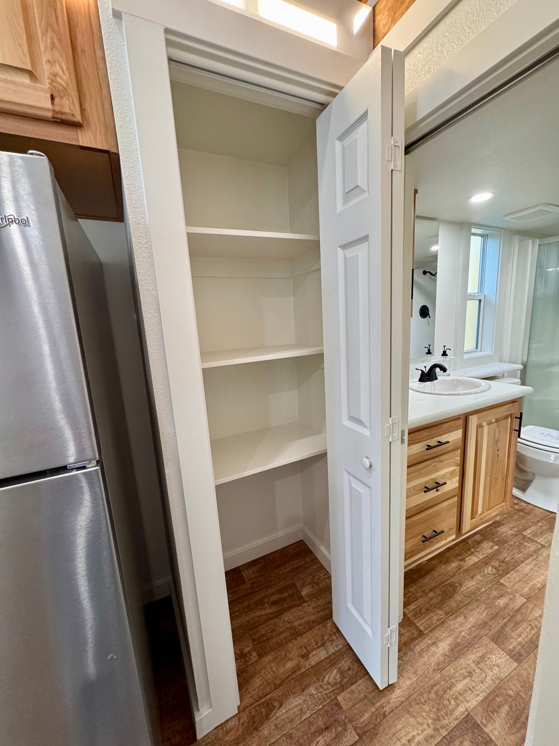 A white floor-to-ceiling pantry with shelves next to a refrigerator and a wooden bathroom vanity.