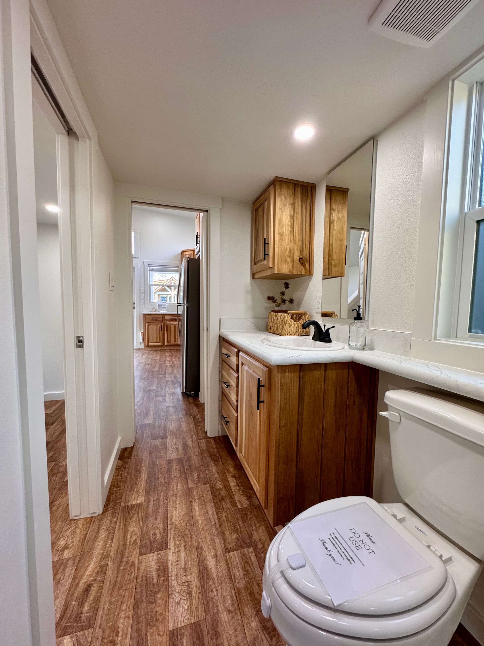 A small bathroom with a wooden vanity, a white toilet, and a doorway looking through to a kitchen with wood floors.