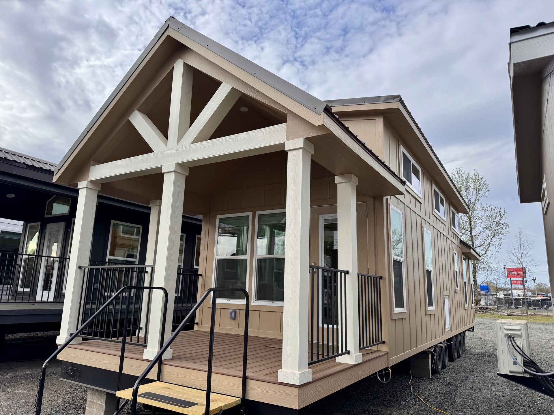Tan tiny house with white trim, porch, and black railing; cloudy sky background.