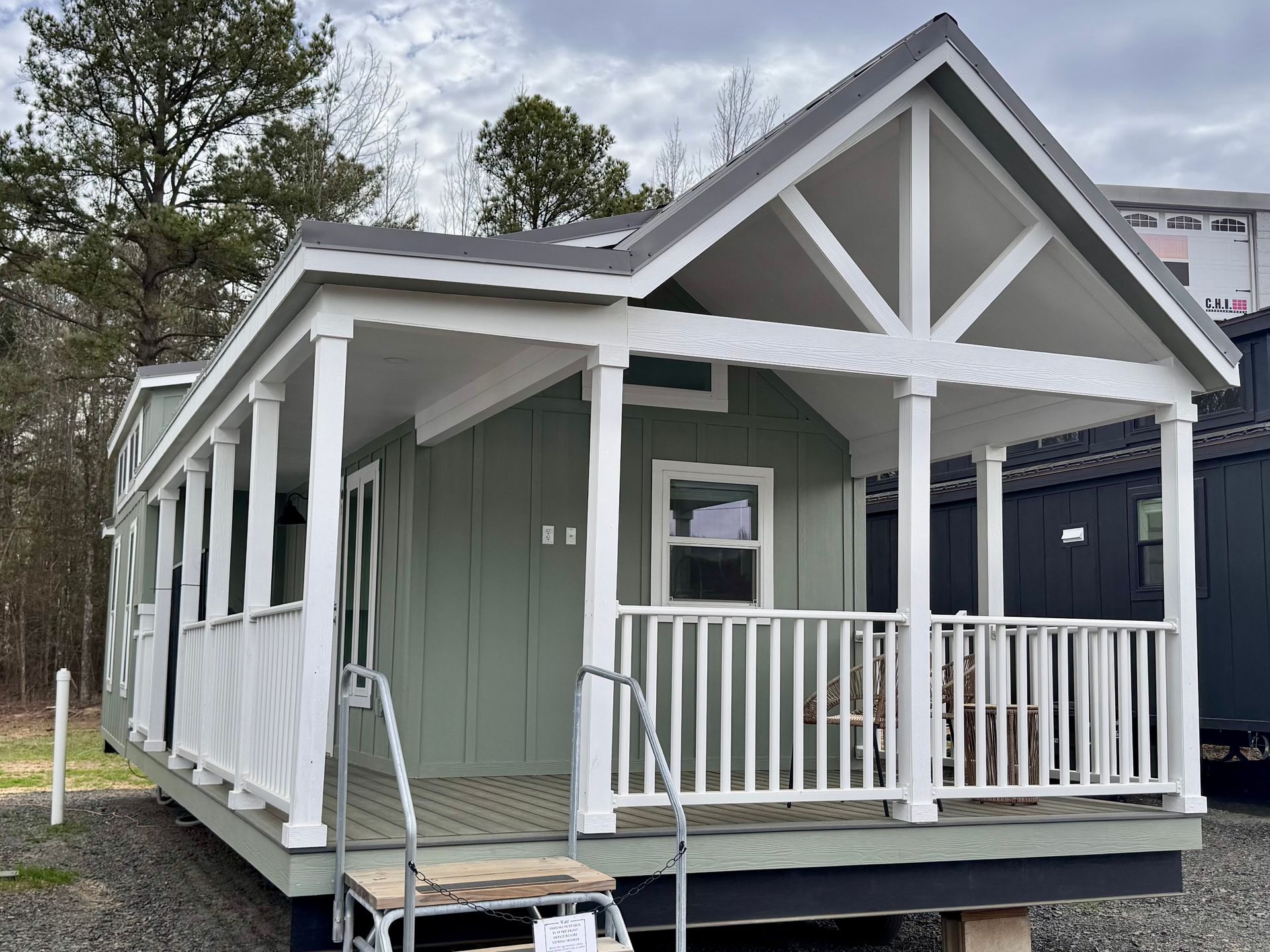 A sage green tiny house with a covered front porch, white railings, and a gabled roof set on a gravel lot.