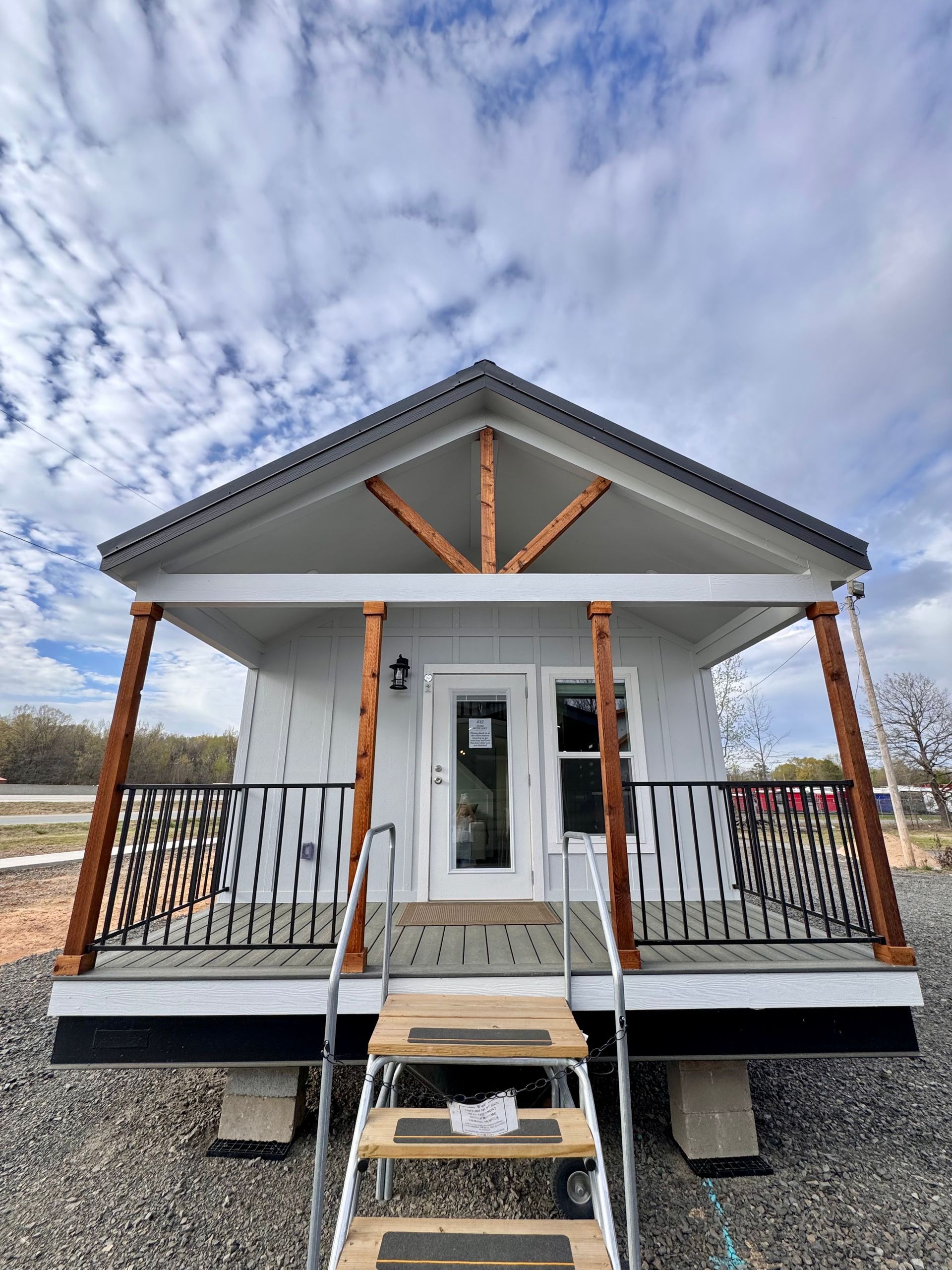 A small, light-gray cabin with a gabled roof, wooden porch pillars, a metal railing, and portable stairs on gravel.
