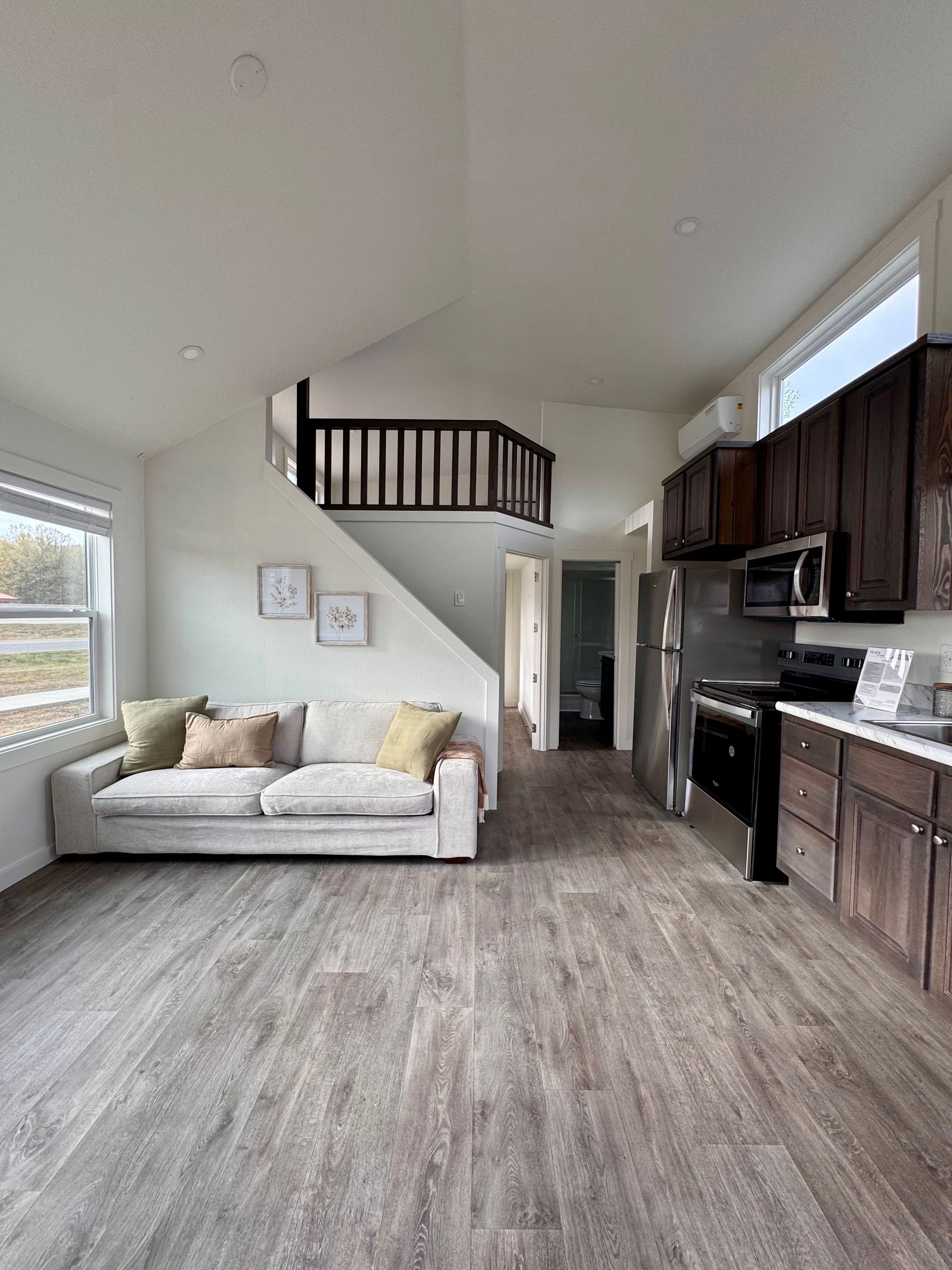 An open-concept studio apartment with a light-colored couch, wood-look flooring, and a kitchen with dark wooden cabinets.