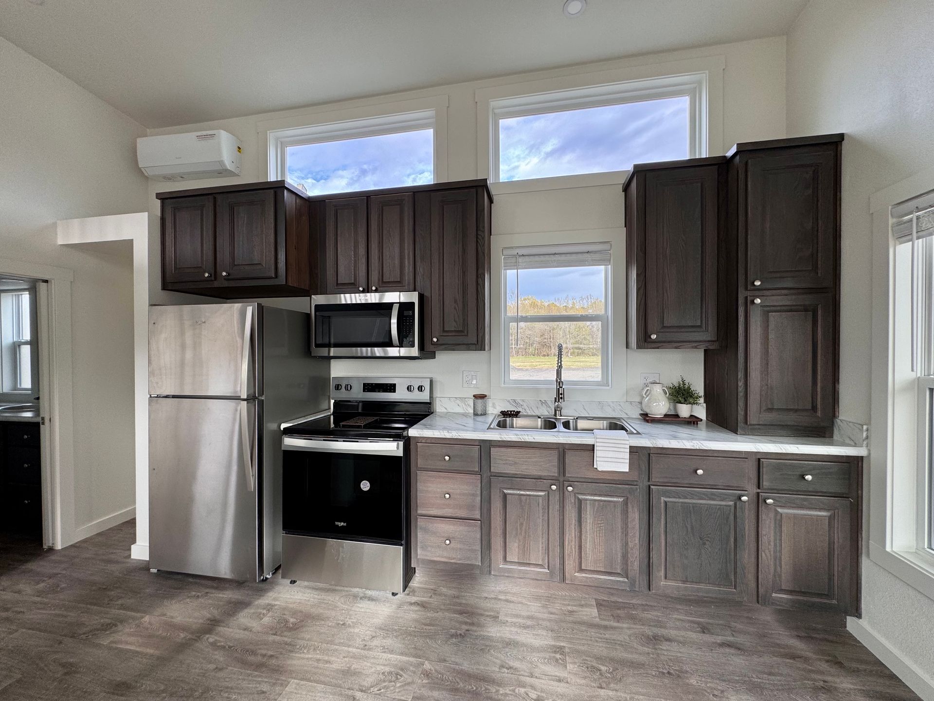 A kitchen with dark wood cabinets, stainless steel appliances, white countertops, and three windows under a slanted roof.