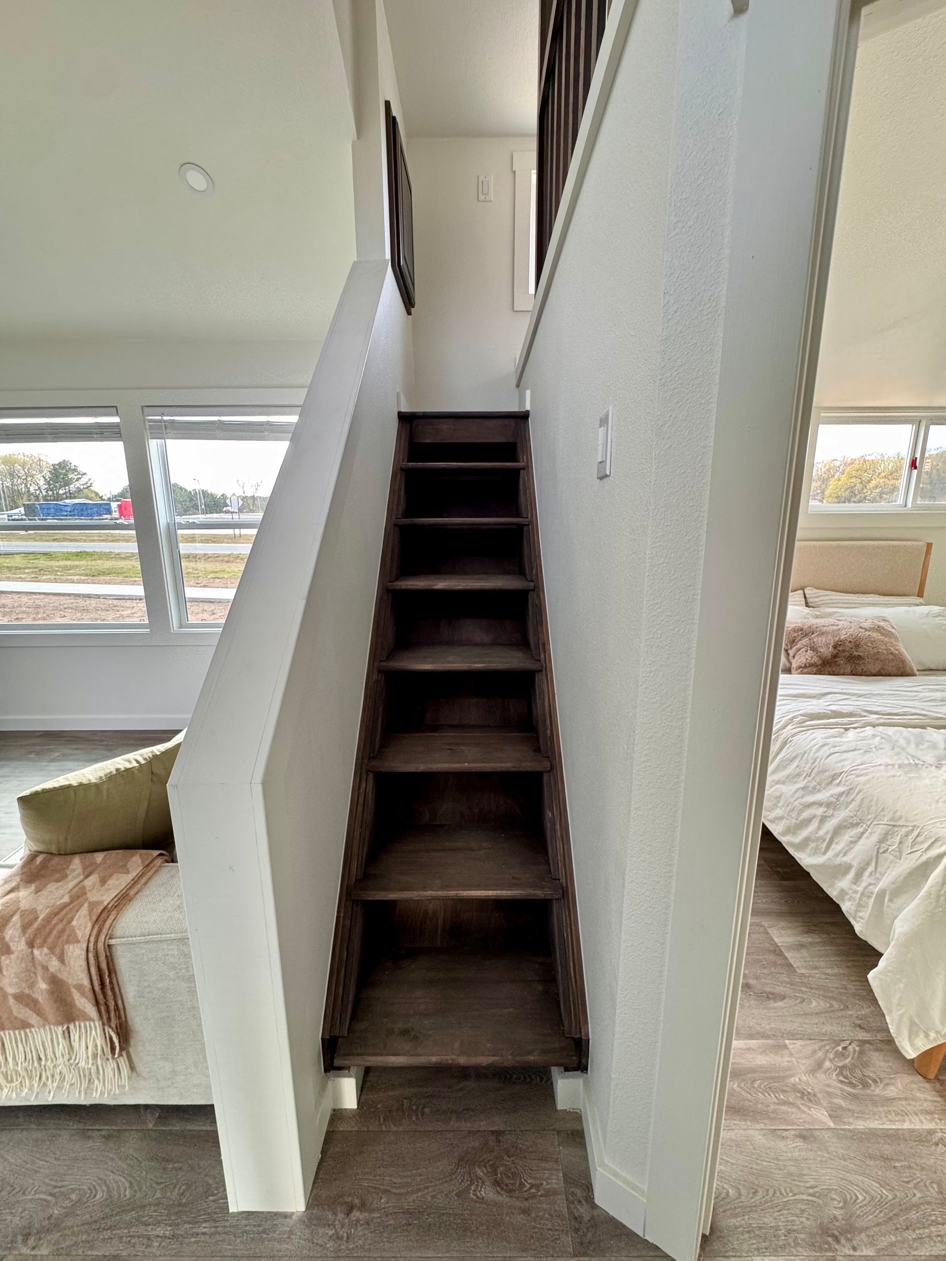 A straight, dark-wood staircase leads upward, centered between a living room and a bedroom in a modern interior.