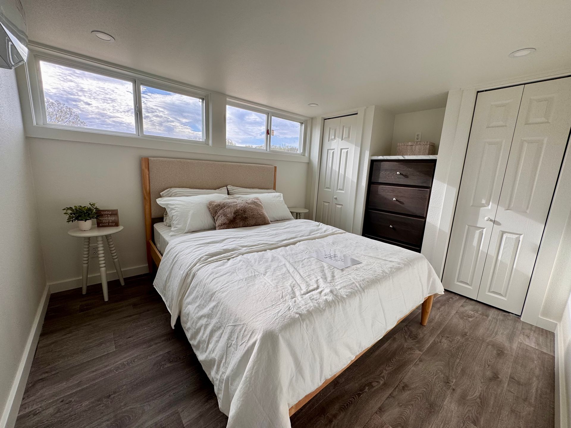 A bedroom with a queen bed, white bedding, dark wood flooring, tall windows, a dark dresser, and white closet doors.