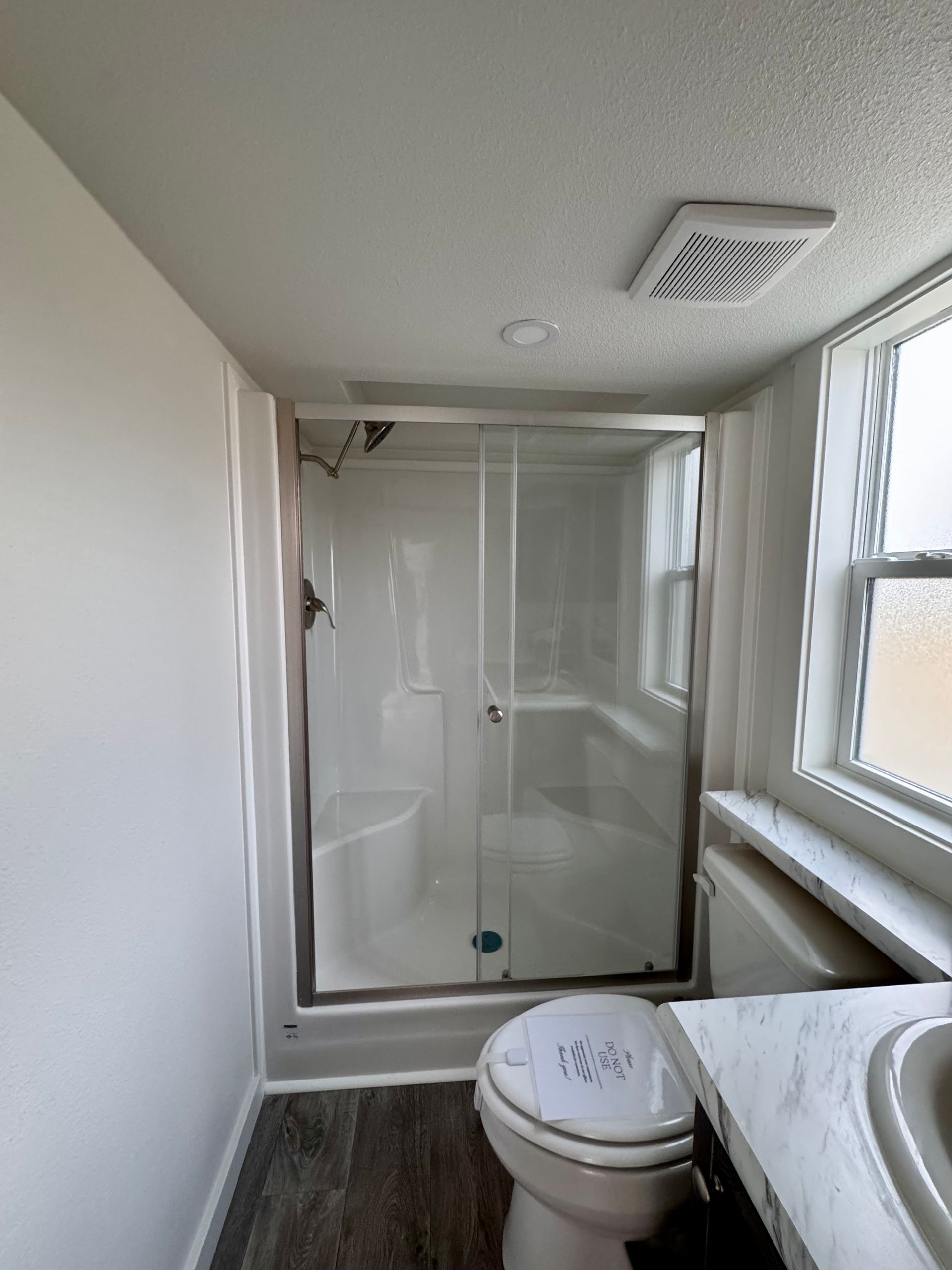 A small bathroom with a glass-enclosed shower stall, a toilet, a vanity counter, and wood-look flooring.