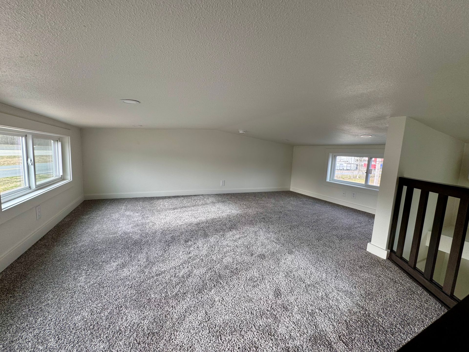 An open attic room with light gray walls, a sloped ceiling, speckled gray carpeting, and a wooden staircase railing.
