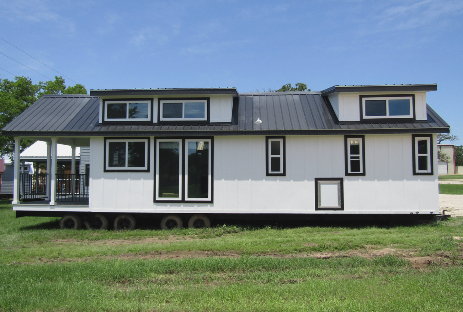 White tiny house on wheels with black accents and a dark roof, in a grassy field.