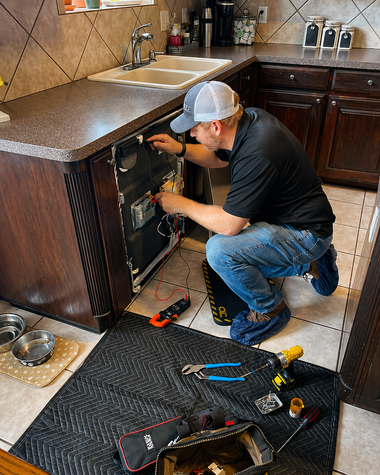 Technician kneeling to repair an appliance in a kitchen, with tools scattered on the floor.