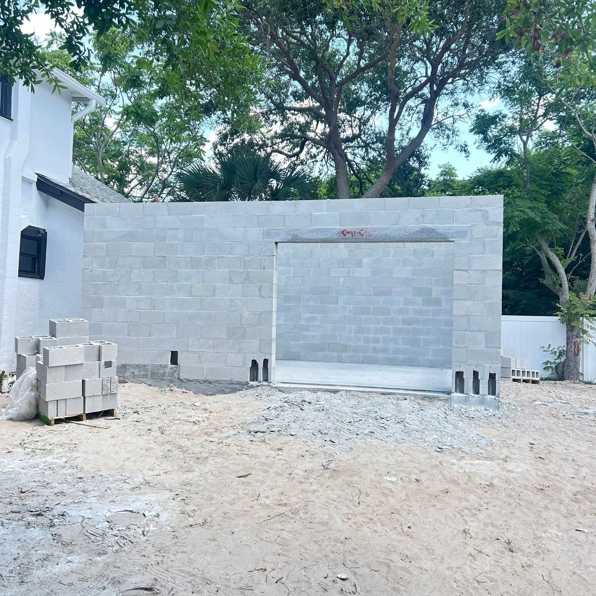 Construction site. Gray cinder block wall with opening, unfinished. White house and trees in background.