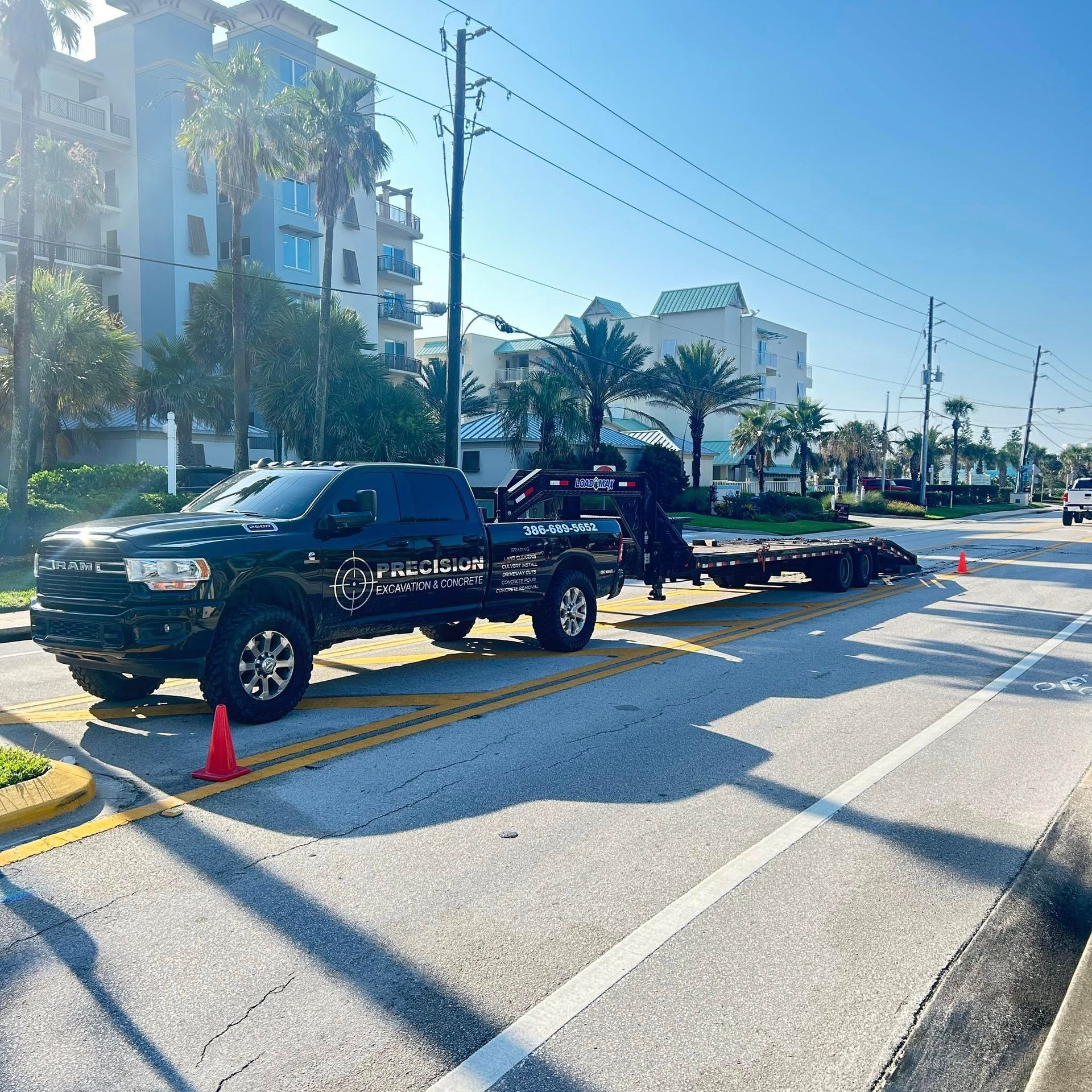 Black pickup truck with trailer on a road, orange cones present. Buildings and palm trees in background.