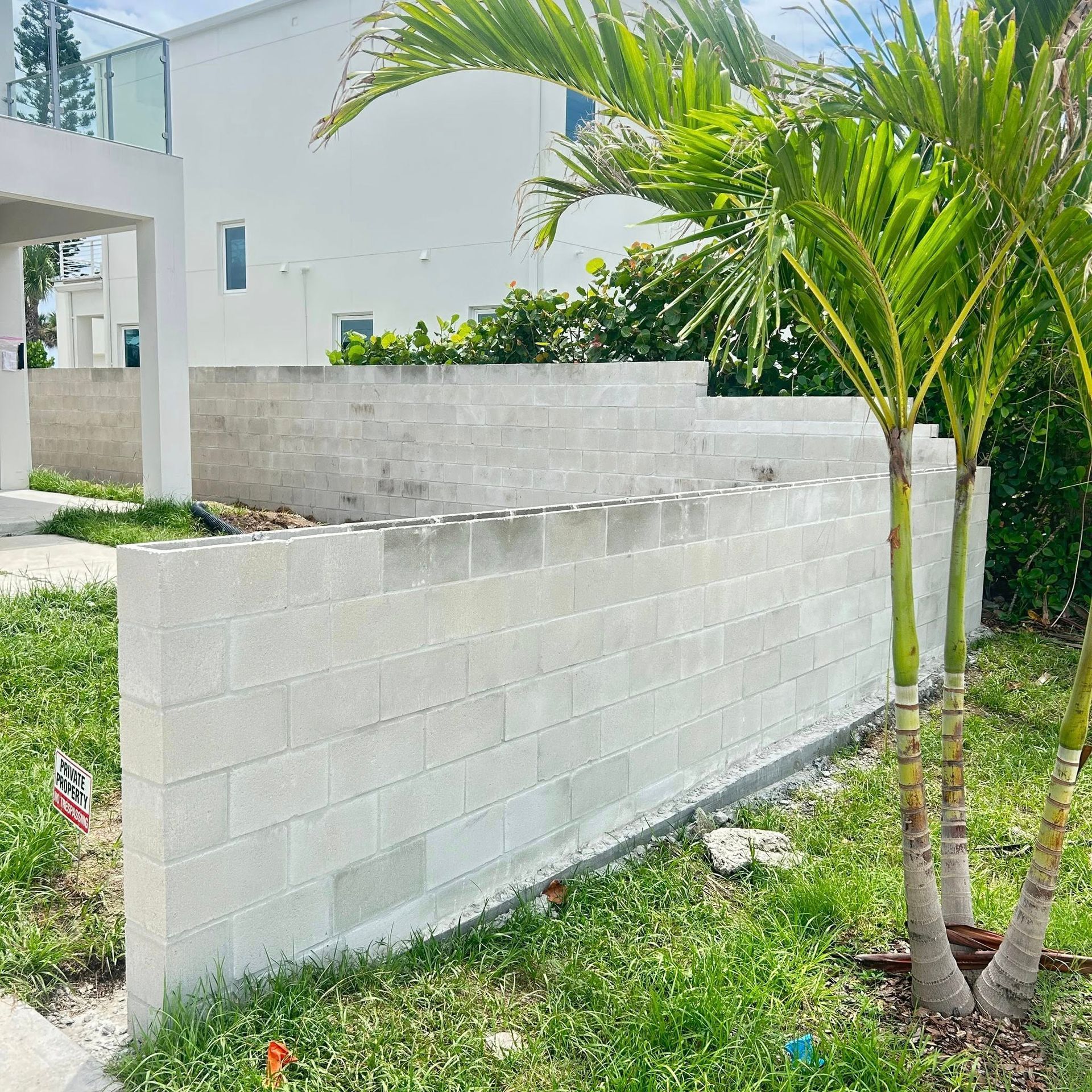 Exterior view: concrete block retaining wall with palms and grass in front of white house.
