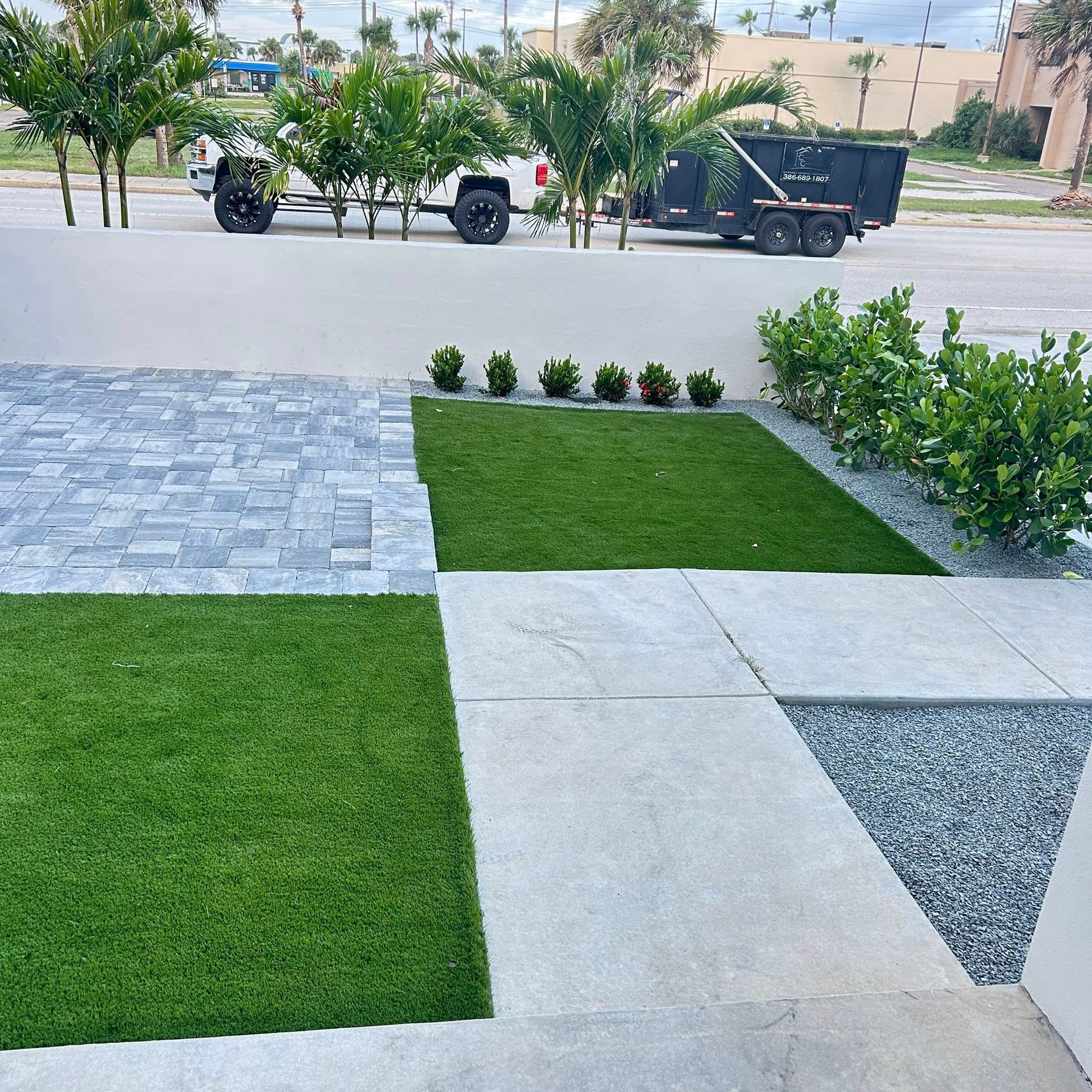 Modern landscaping with pavers, artificial turf, and a small green shrub border with a white wall and a truck in the background.
