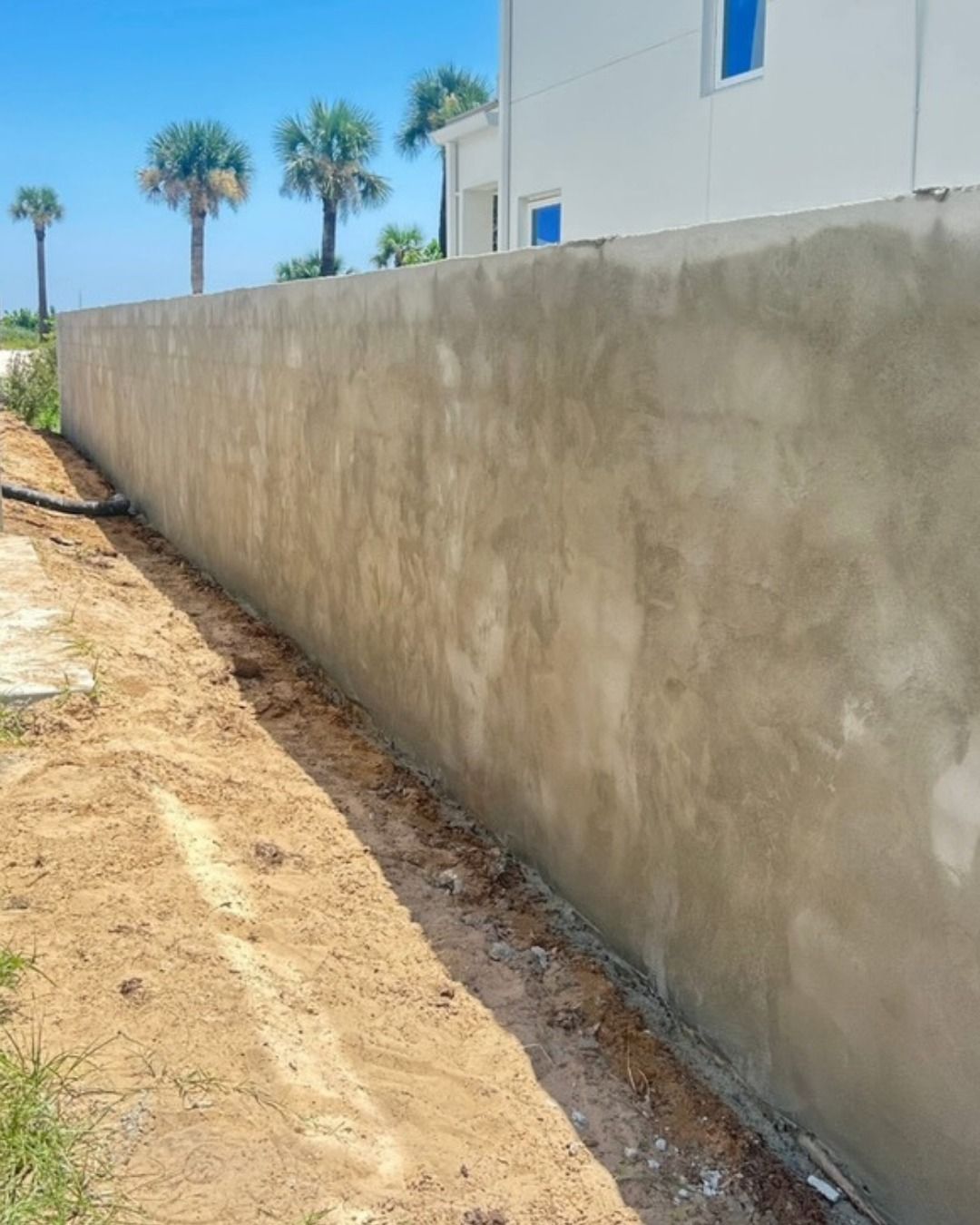 Concrete wall along a dirt path, near a white building and palm trees under a blue sky.