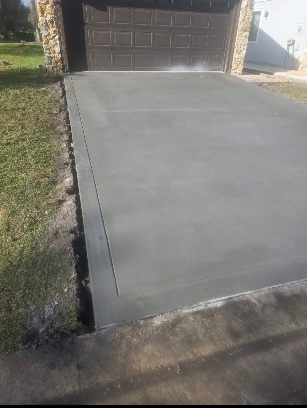 Freshly poured gray concrete driveway with a garage in the background, bordered by grass and a road.