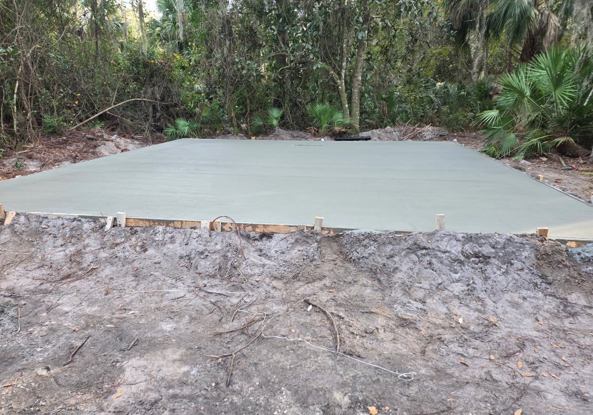 Newly poured concrete slab in an outdoor setting, surrounded by dirt and vegetation.