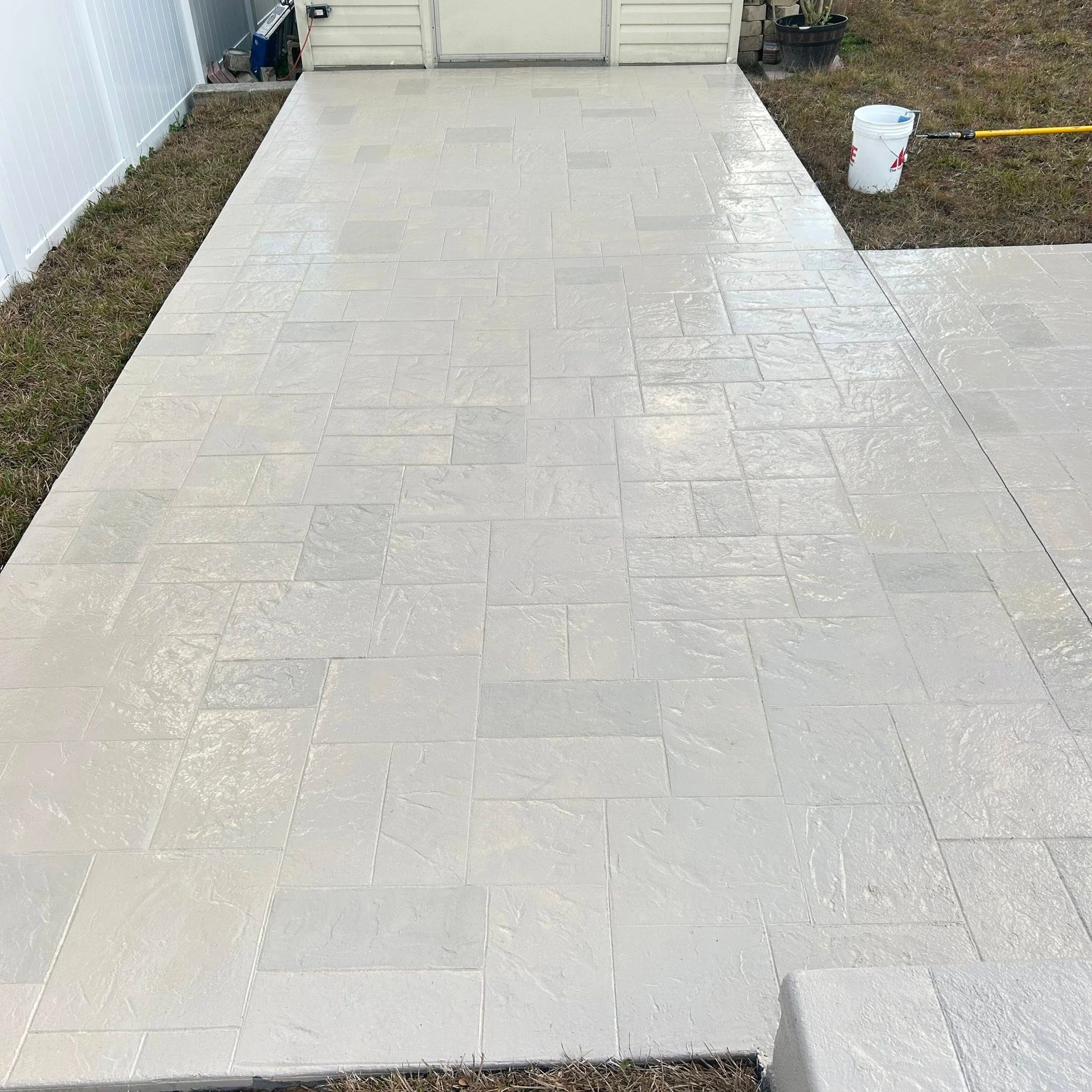 Gray stamped concrete patio with rectangular and square patterns; next to a white fence and grass.