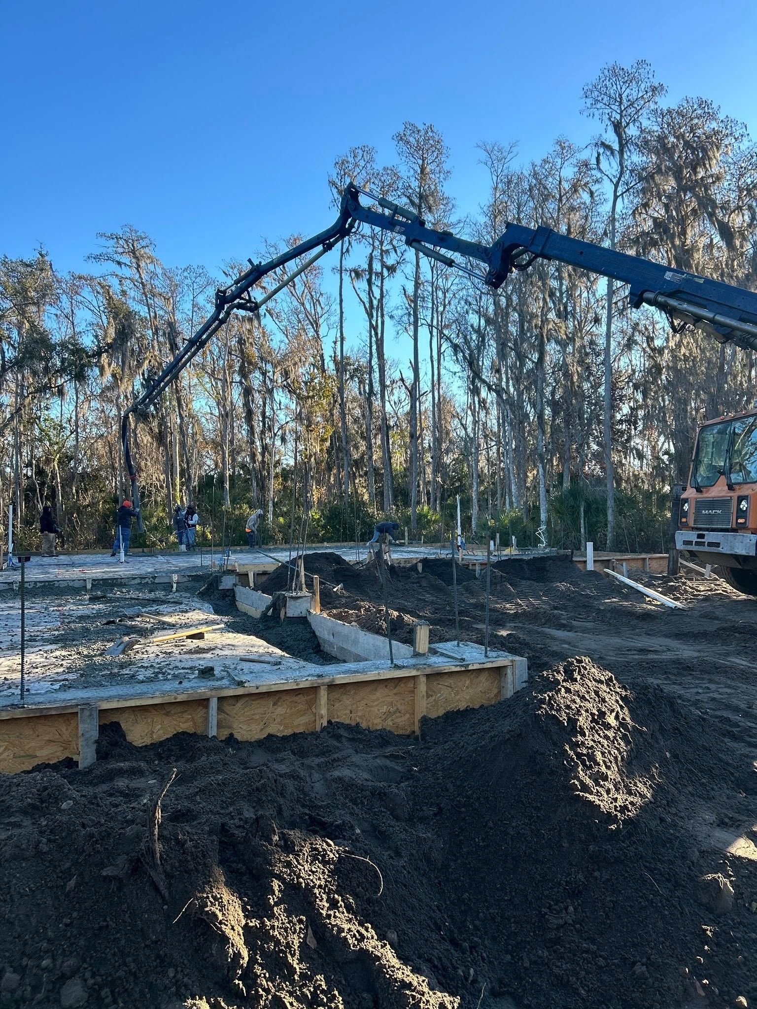 Construction site with concrete being poured for a foundation; pump truck, workers, blue sky, trees.