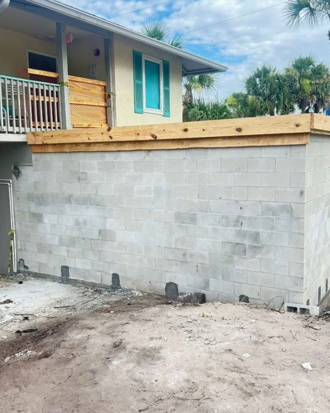 Concrete block wall under a house with new wooden beams on top, sandy ground.