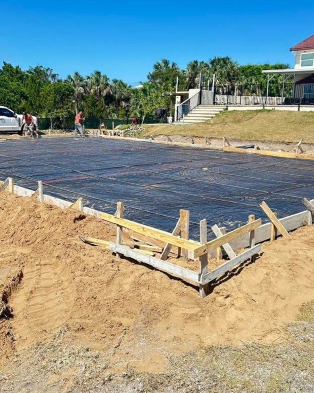 Construction site: concrete foundation with rebar, wooden forms, and workers in sunny outdoor setting.