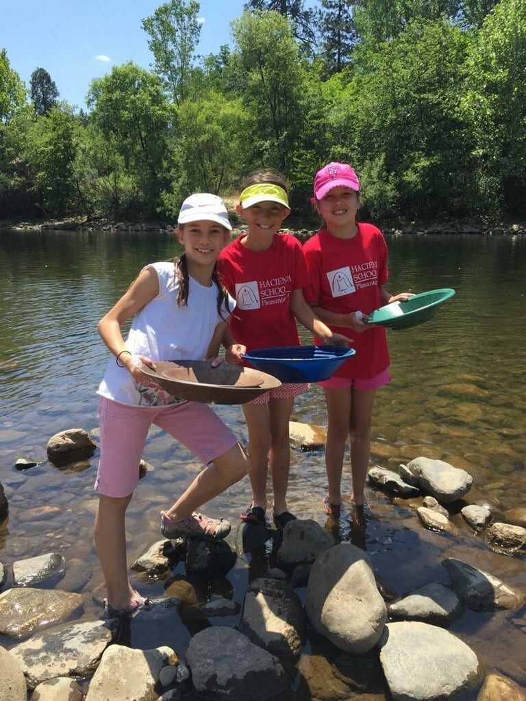 Three smiling elementary students stand in a rocky riverbed holding colorful pans during a hands-on history field trip.